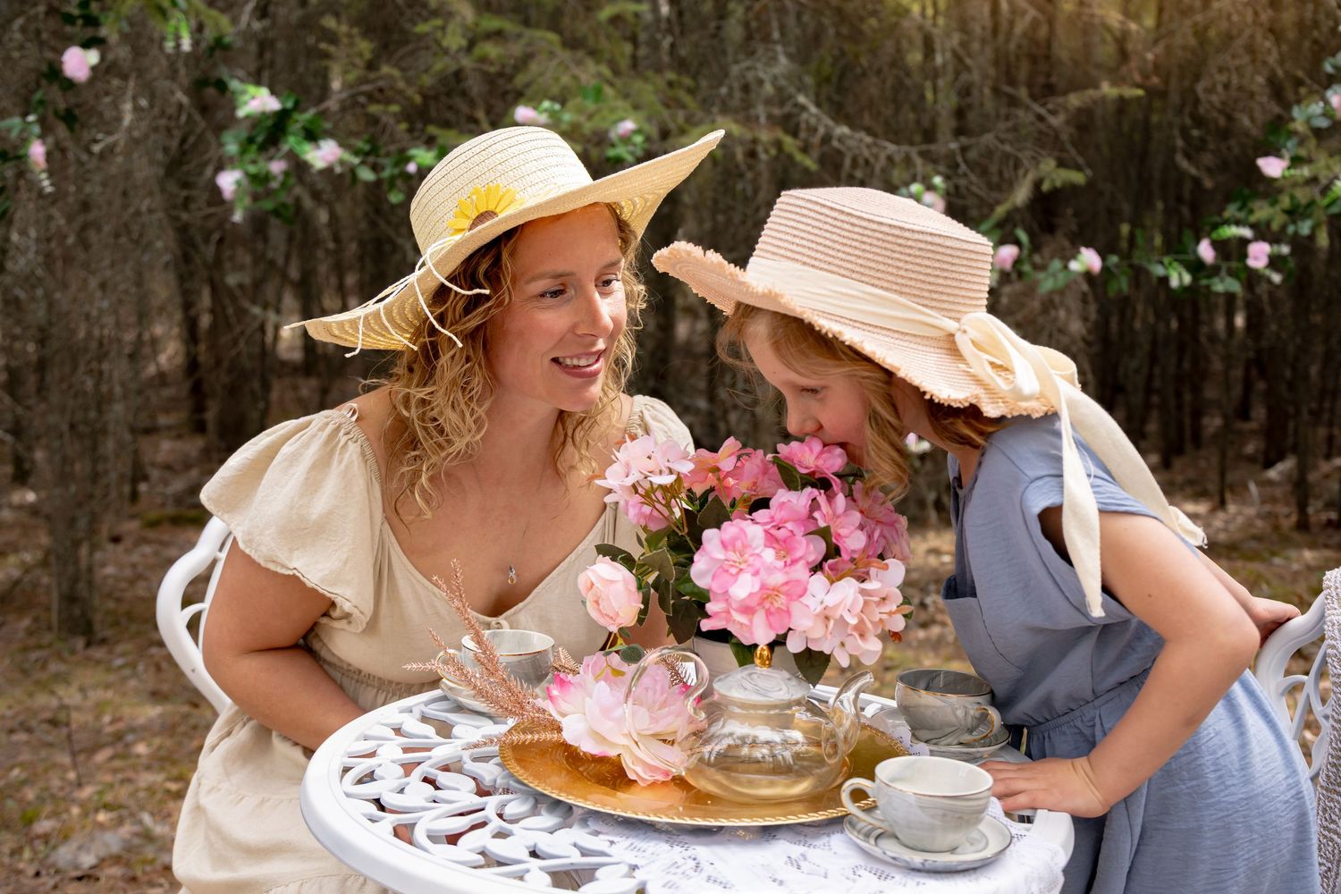 Two women in sun hats enjoy a floral tea party outdoors, smiling over pink roses at a white garden table.