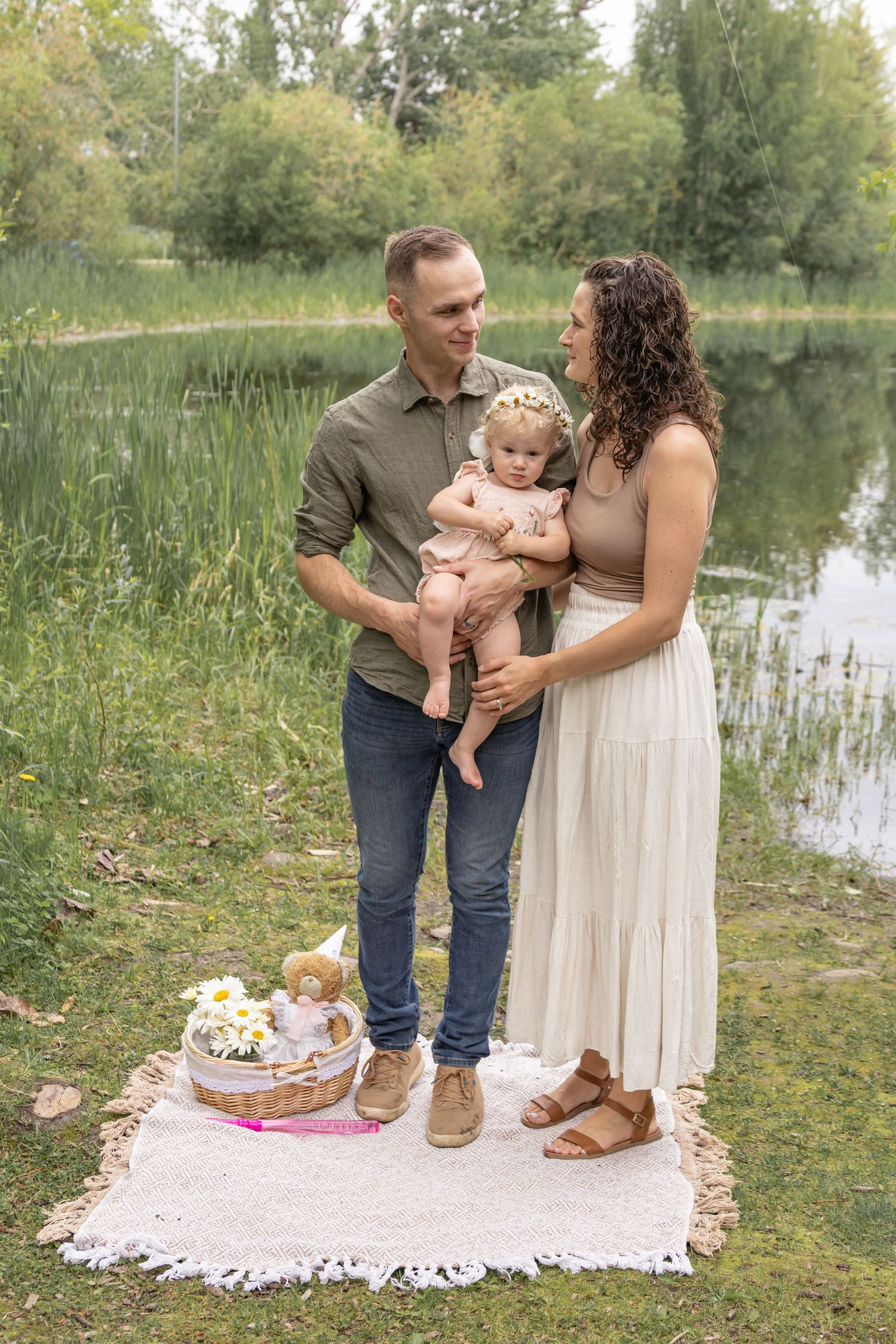 A man and woman hold their baby girl on a white picnic blanket by a calm pond, with a flower-filled basket and teddy bear nearby.