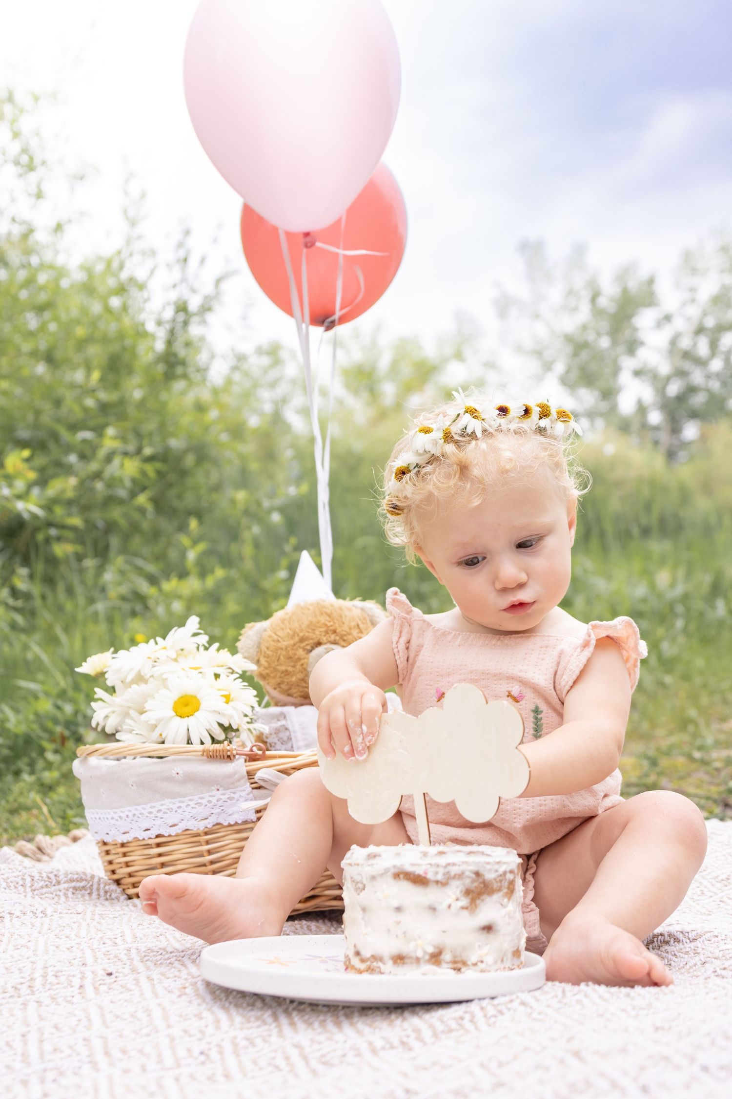 Baby girl in a daisy flower crown sits on a blanket with a smash cake, pink balloons, and a daisy-filled wicker basket outdoors.
