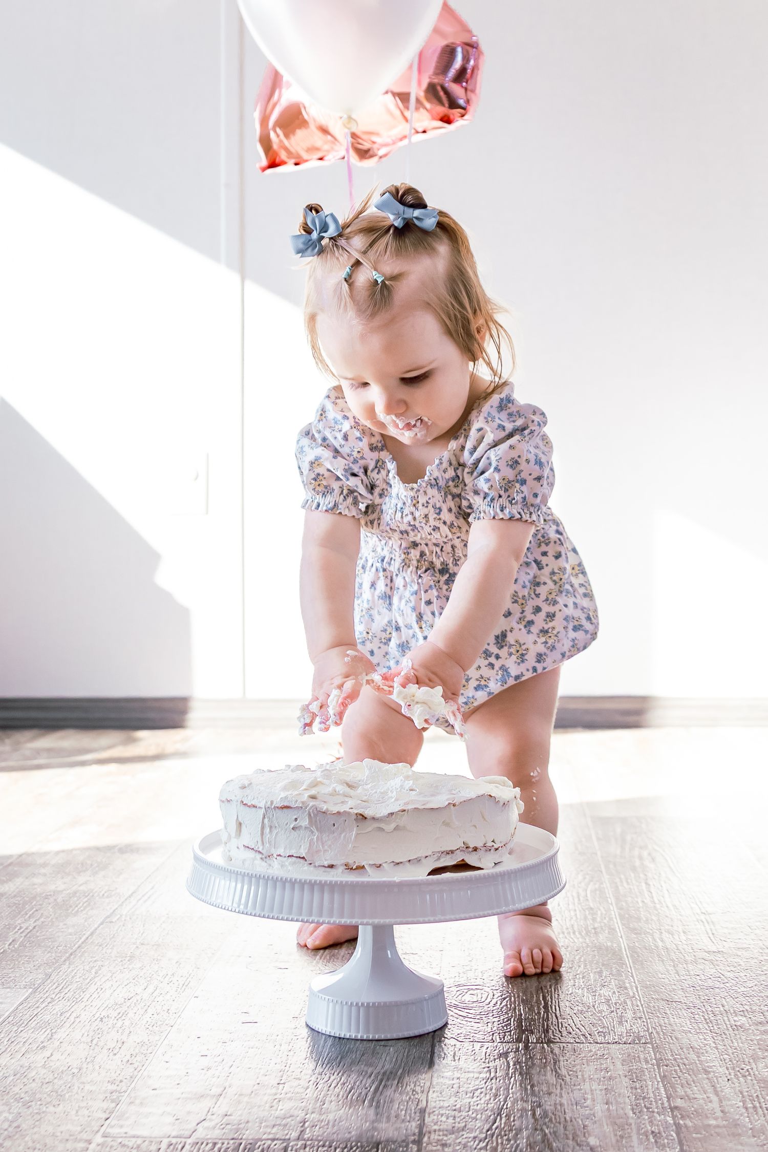 Toddler girl in floral dress smashing white birthday cake on stand, with pink balloons in background.