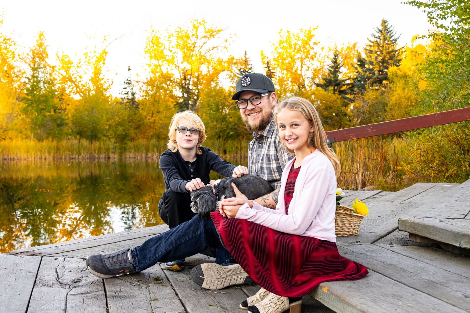 Father sits on wooden dock with two young children amid vibrant autumn foliage by a lake.