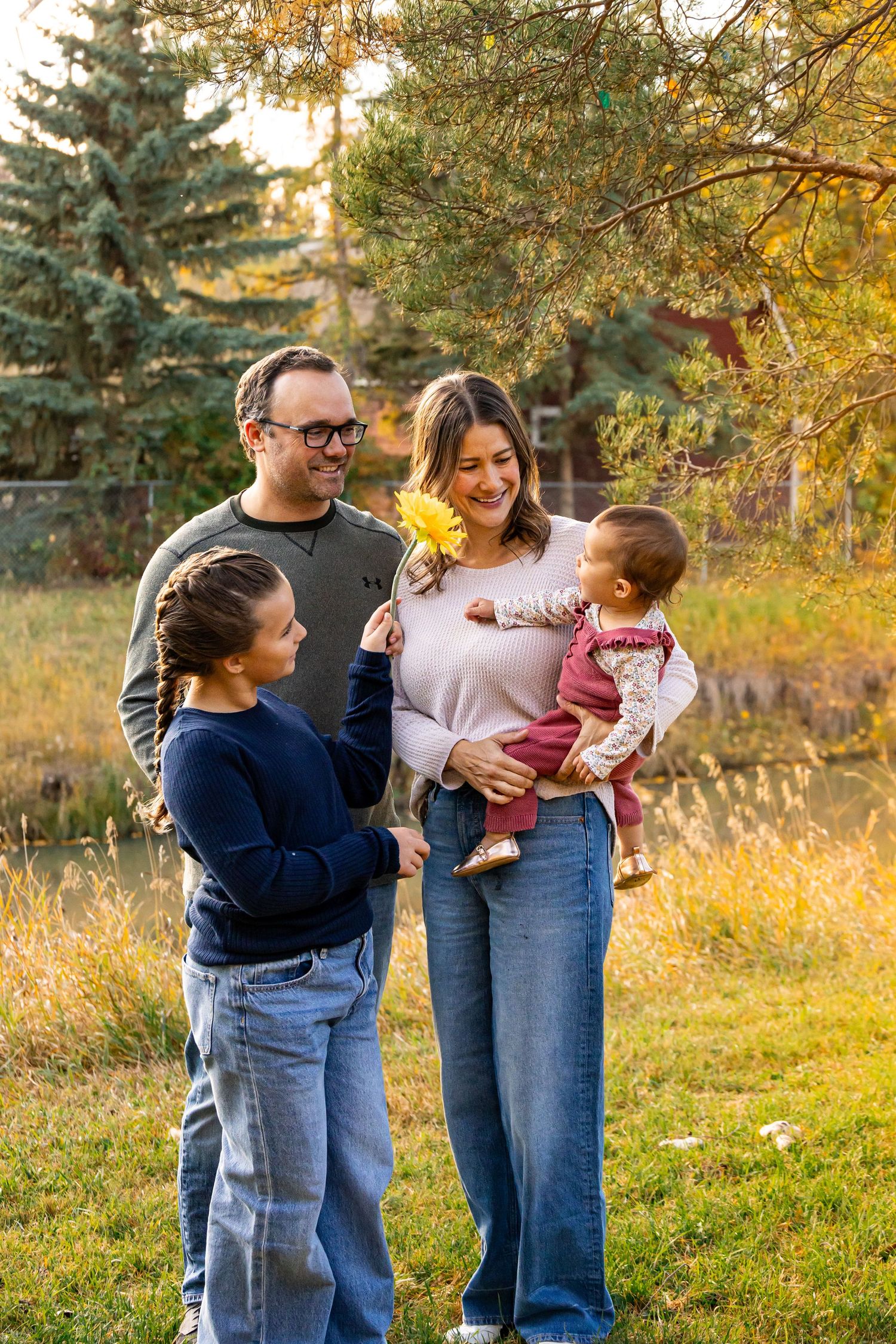 Family of four enjoying autumn outdoors, parents smiling with two children, holding yellow flowers in a park setting.