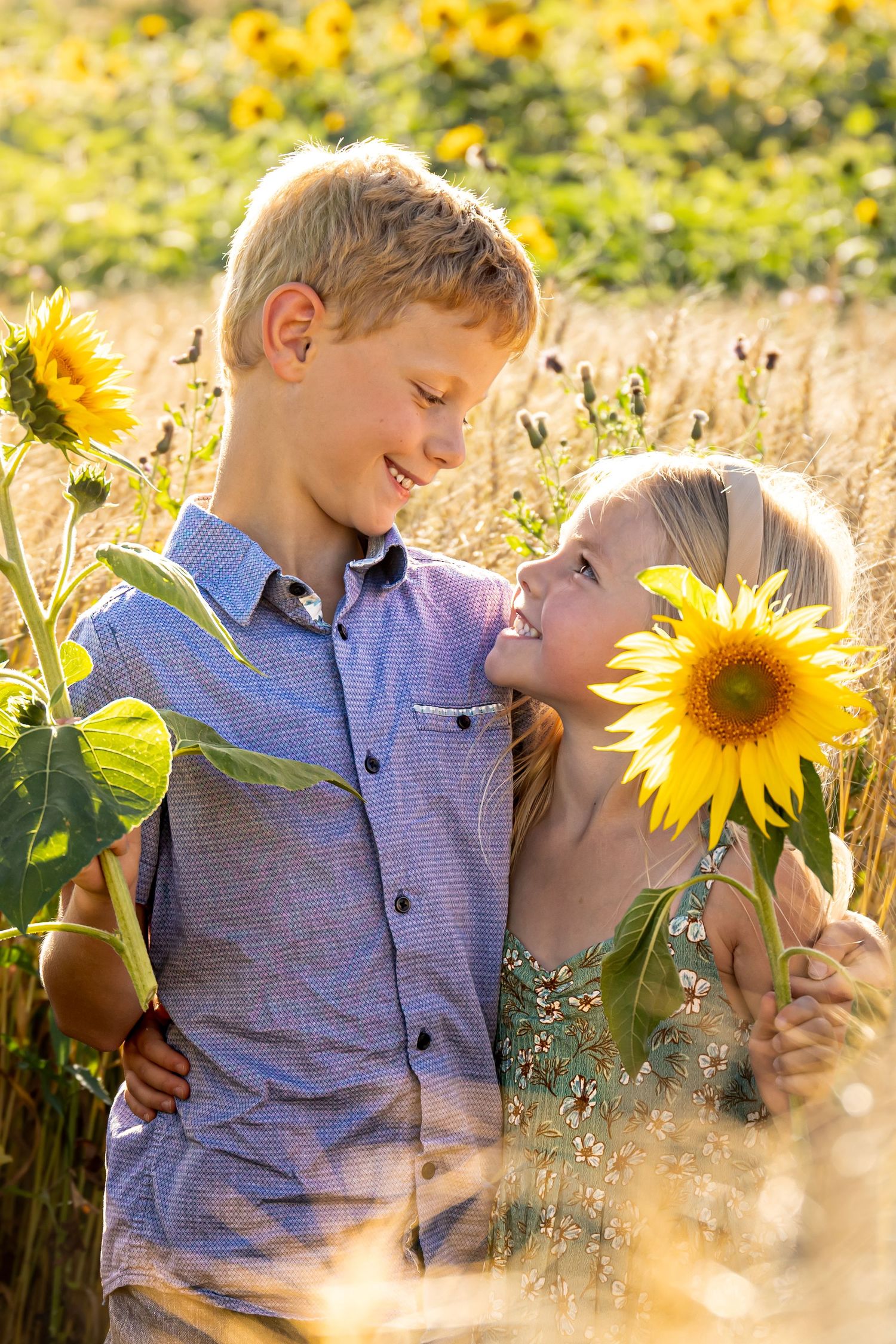 Two happy children smiling at each other in a sunny sunflower field, holding bright yellow sunflowers.