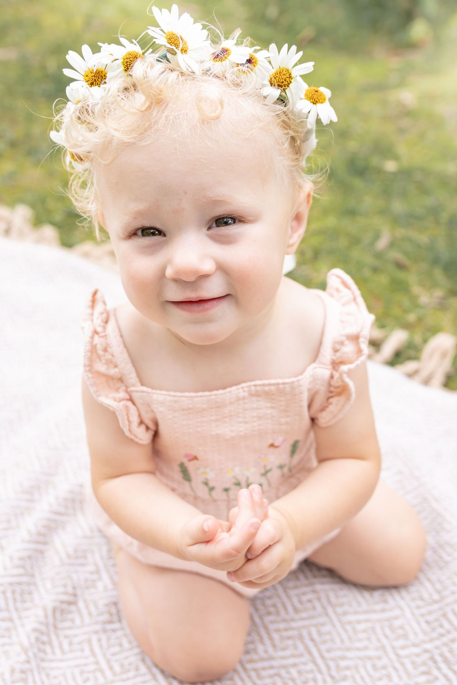 Smiling toddler with curly blonde hair and a daisy flower crown kneeling on a soft blanket in a grassy park, wearing a pink ruffled romper.