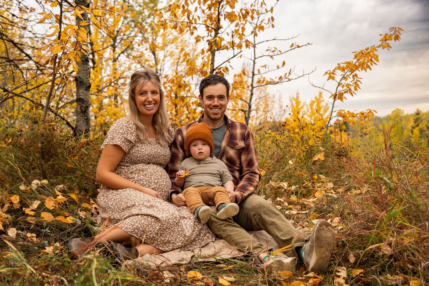 Pregnant woman and man sit outdoors with toddler among golden autumn leaves and trees for a fall family portrait.