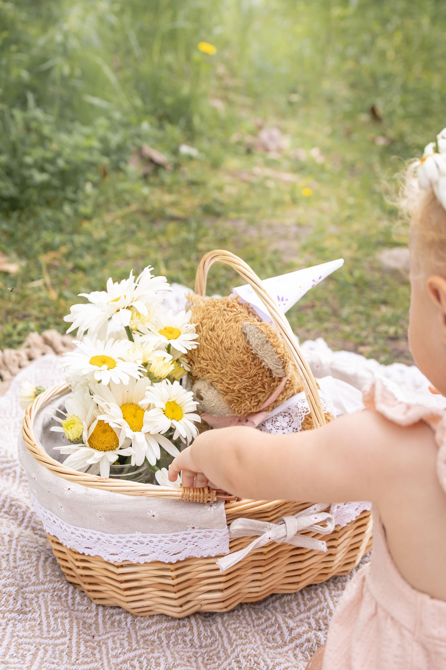 Toddler in a pink outfit reaches into a wicker basket with white daisies and a teddy bear in a party hat on a blanket outdoors.