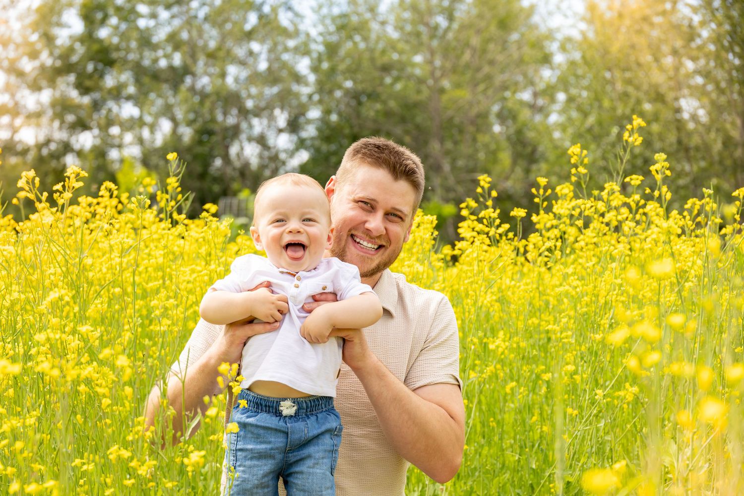A smiling man holds a laughing baby in a sunny field of yellow wildflowers with green trees in the background.
