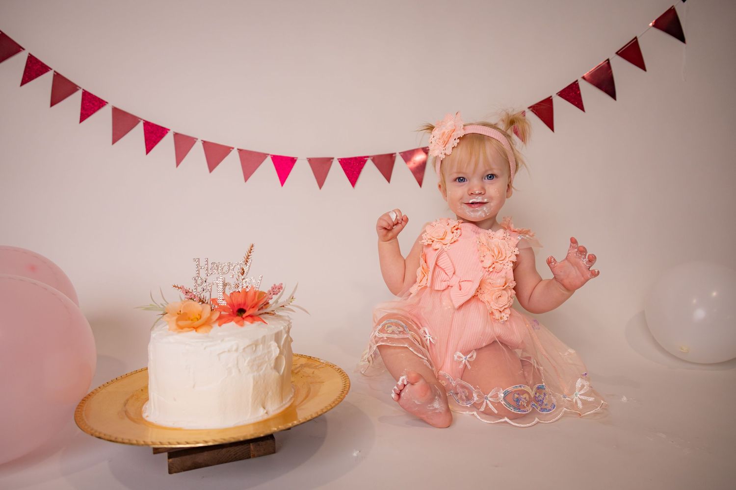 Baby girl in a pink tutu dress and floral headband sits beside a white smash cake with orange flowers at a first birthday photoshoot.