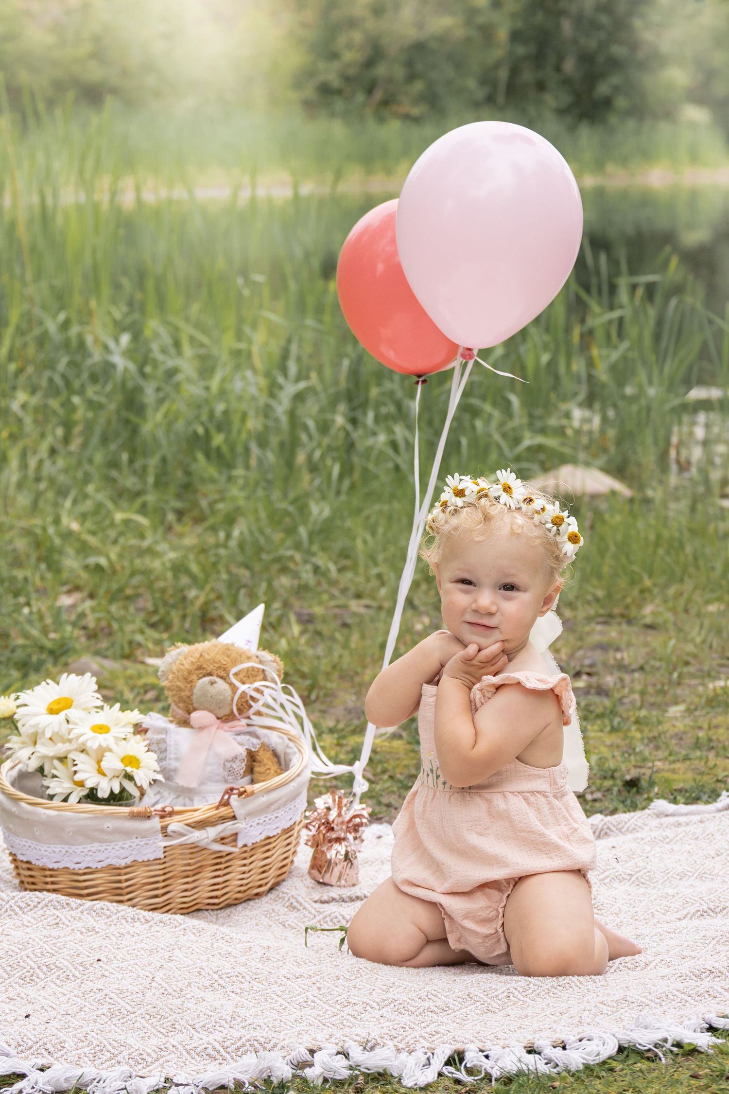 Toddler in a pink romper and daisy flower crown sits on a white blanket near pink balloons and a teddy bear in a wicker basket.