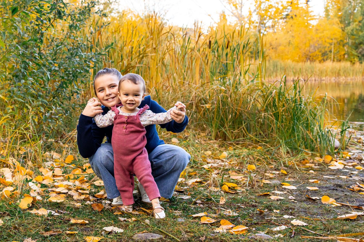 Mother and toddler smiling together in autumn park with colorful yellow leaves and pond in background.