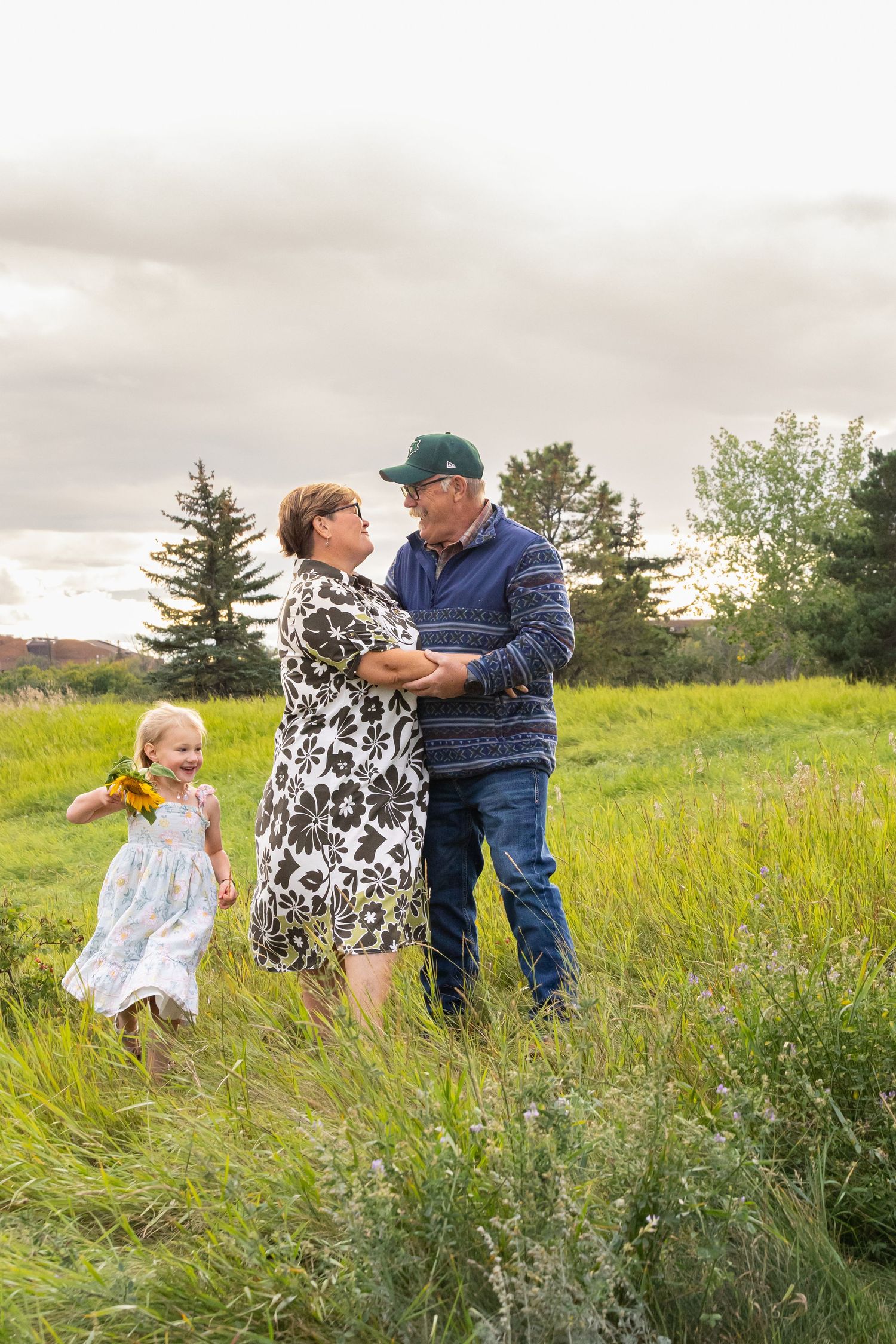 Grandparents dancing in a green field while a young girl in a white dress holds sunflowers on a cloudy day.