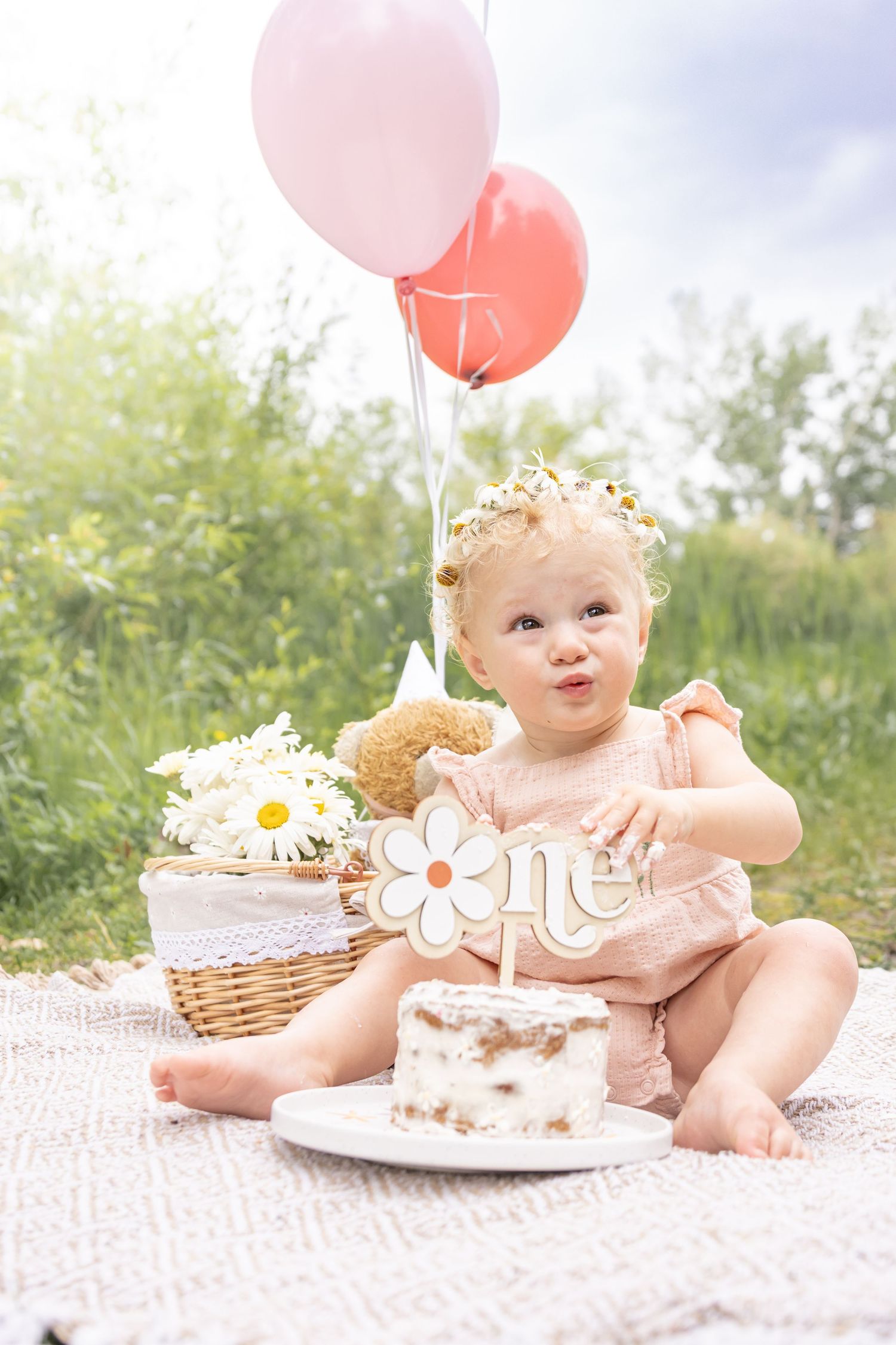 Toddler in a pink romper and flower crown sits on a blanket with a smash cake, pink balloons and daisies for a first birthday photo.