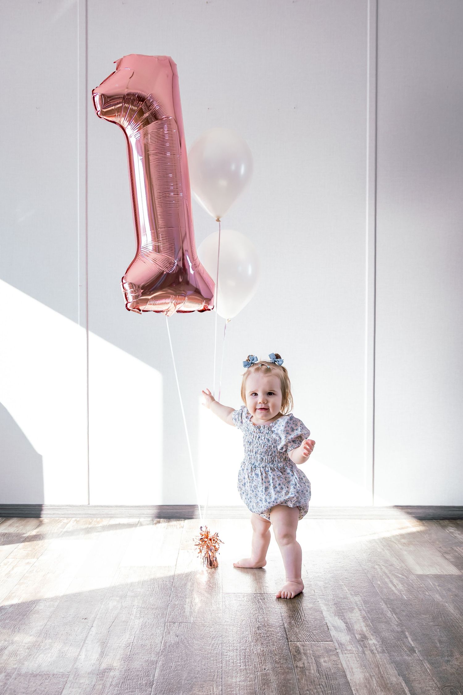 Toddler in a floral romper and bow headband stands barefoot holding a rose gold number one balloon and white balloons for a first birthday.