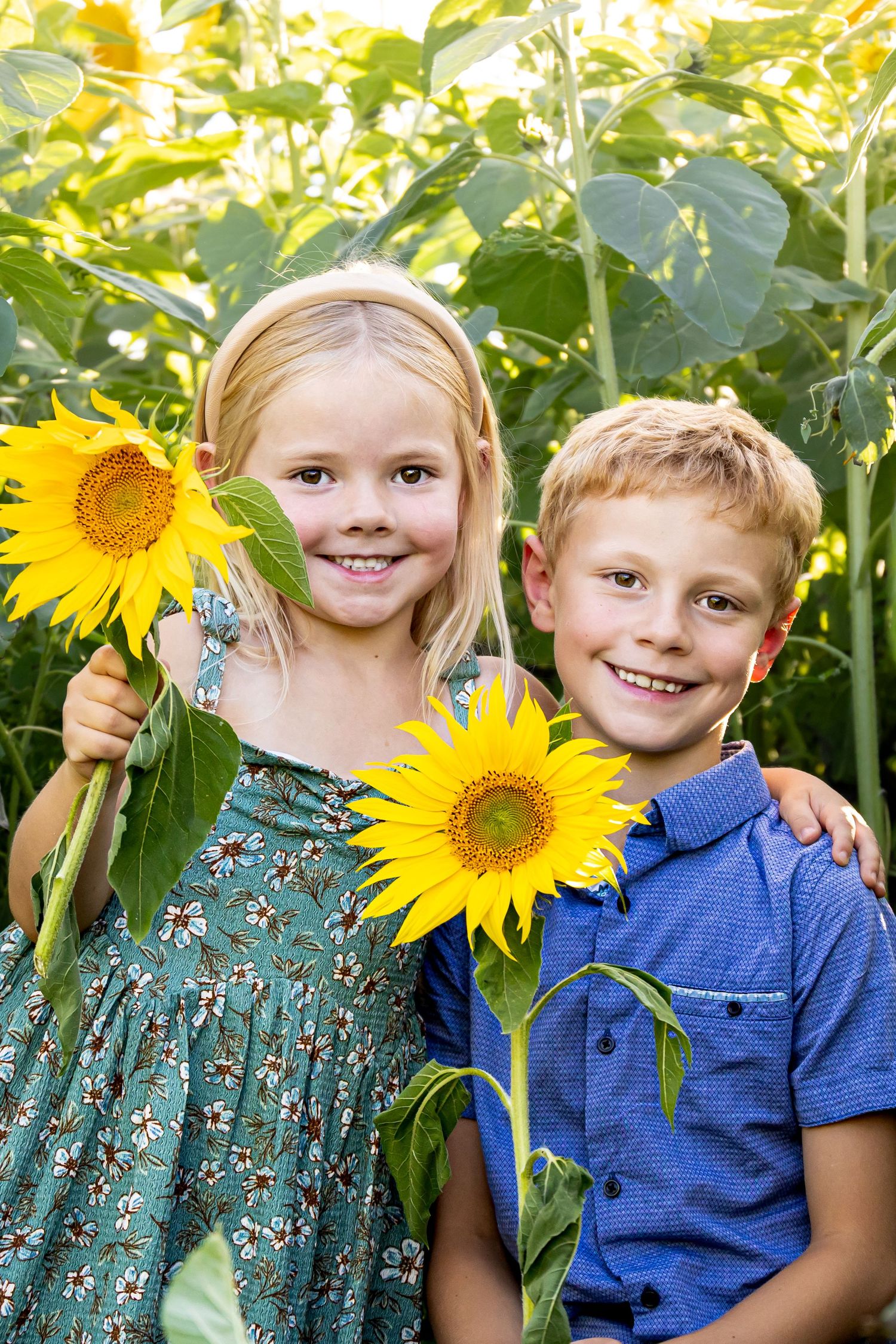 Two smiling children holding bright yellow sunflowers in a lush sunflower field, girl in floral dress and boy in blue shirt.