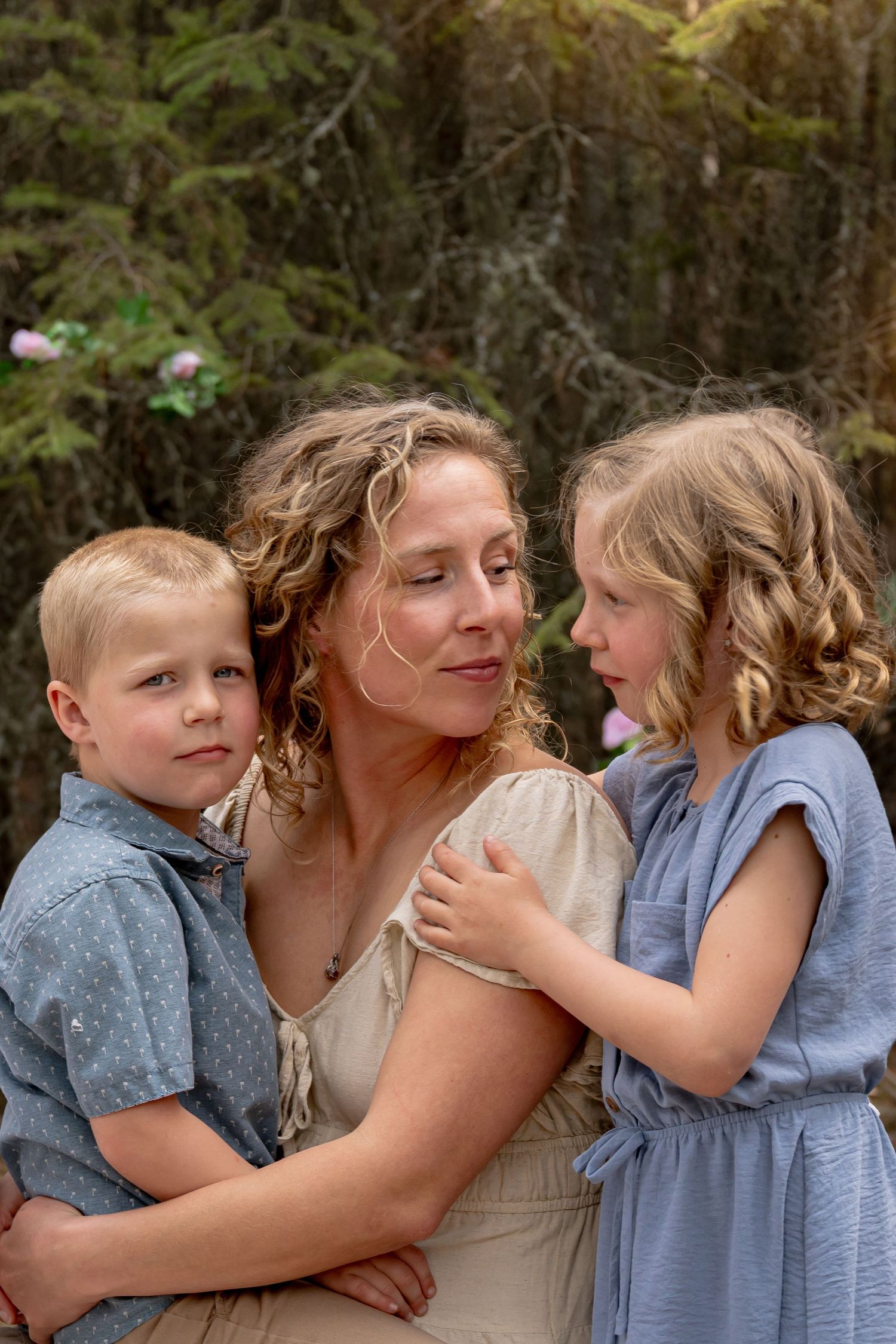 A mother with curly blonde hair embraces her two young children outdoors among lush green trees.
