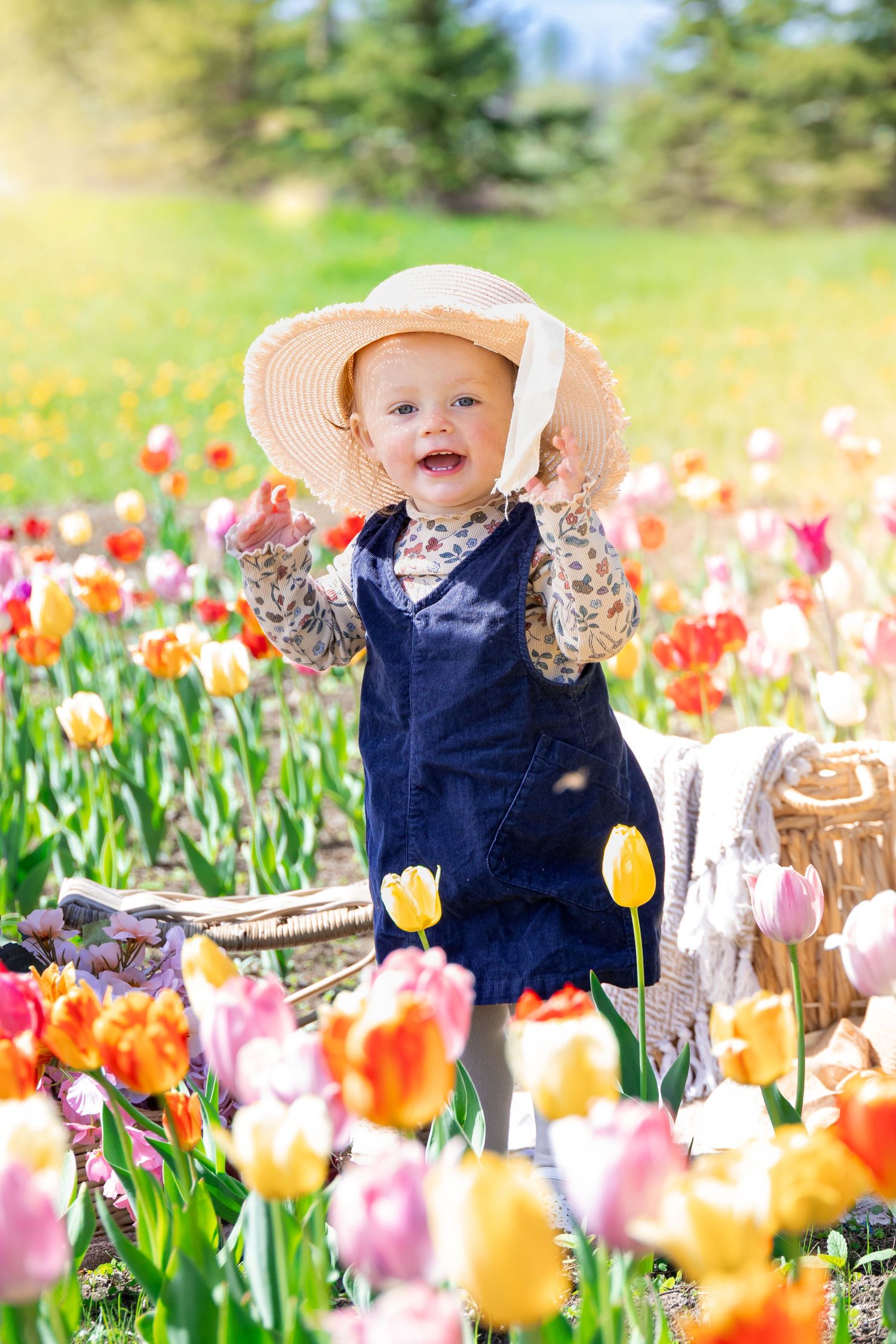 Smiling baby in straw hat and denim overalls standing among colorful tulips in a sunny spring garden.