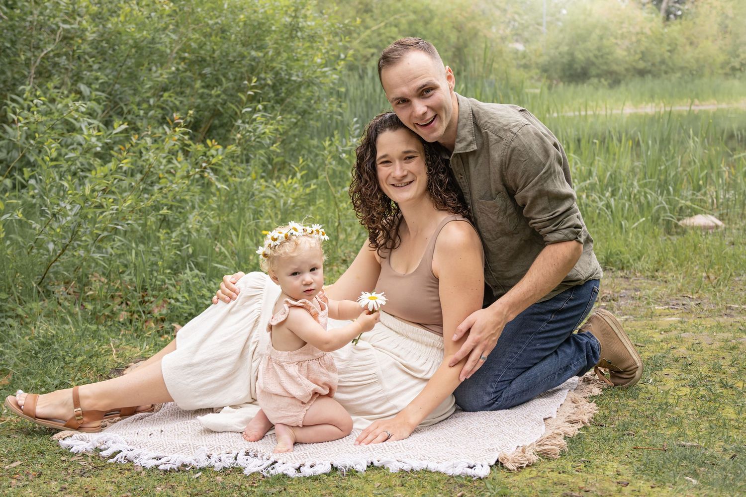 Smiling family of three posing on a cream blanket in a green meadow, with a toddler in a flower crown sitting between her parents.