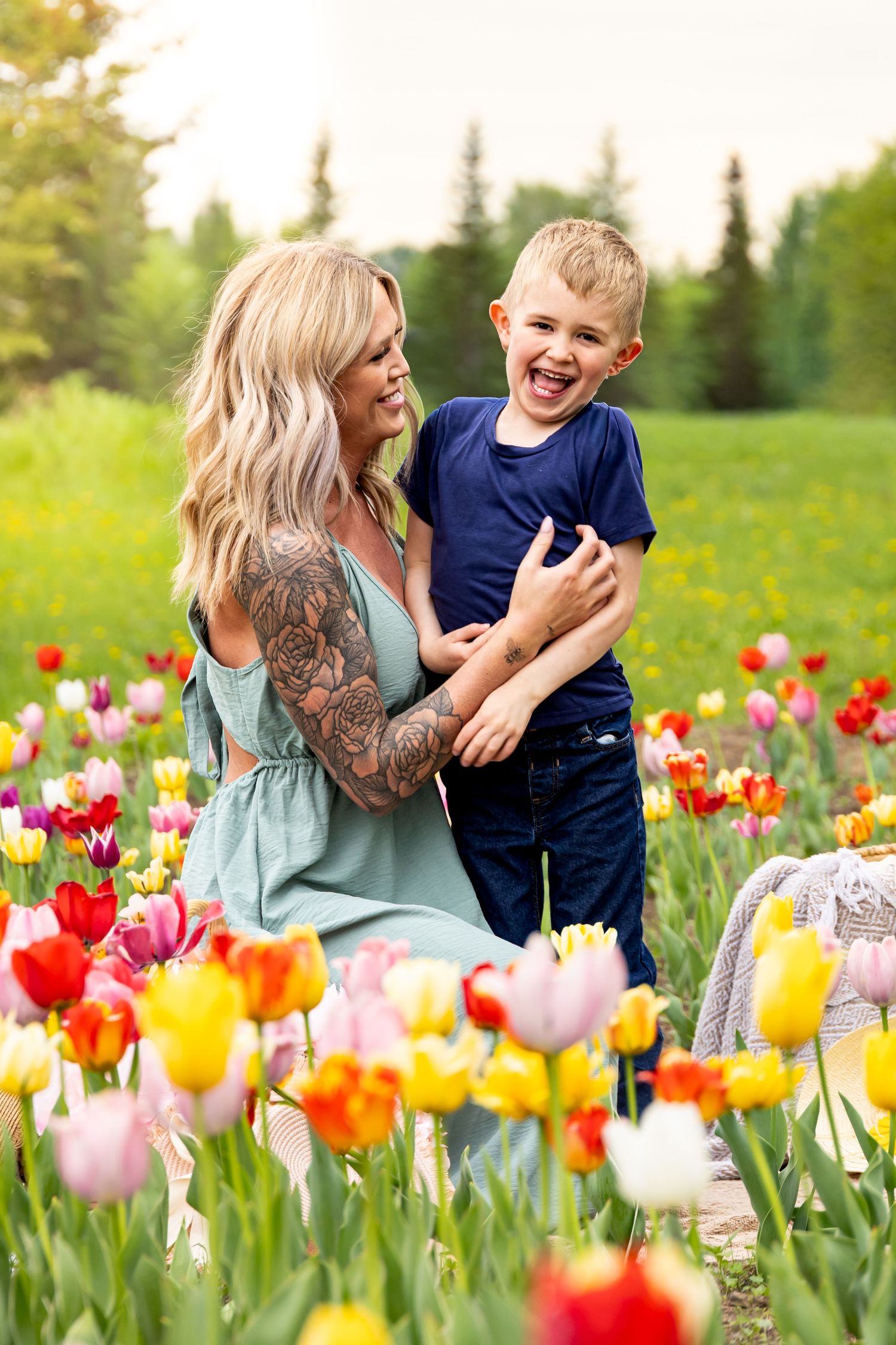 A tattooed blonde mother laughs with her young son in a vibrant tulip field with colorful red, yellow, and pink blooms.