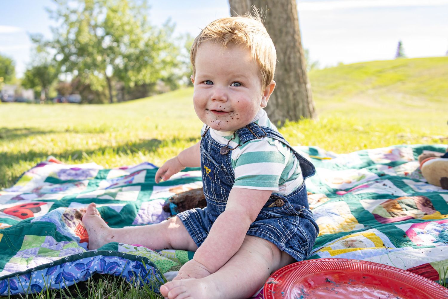 Happy toddler sitting on a colorful quilt outdoors in a sunny park wearing denim overalls and a striped shirt.
