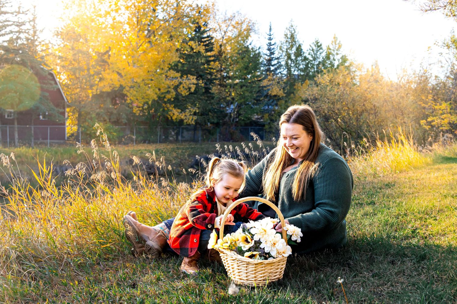Woman and toddler sitting in golden autumn grass with a flower basket, smiling outdoors near trees.