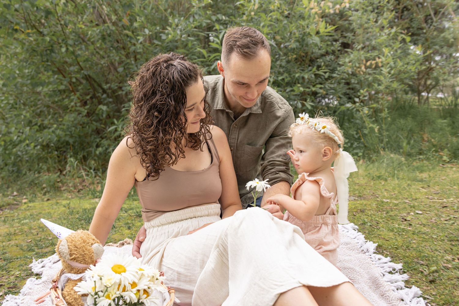 Smiling family of three enjoying a sunny outdoor picnic on a lace blanket, with a baby in a flower crown and fresh daisies nearby.
