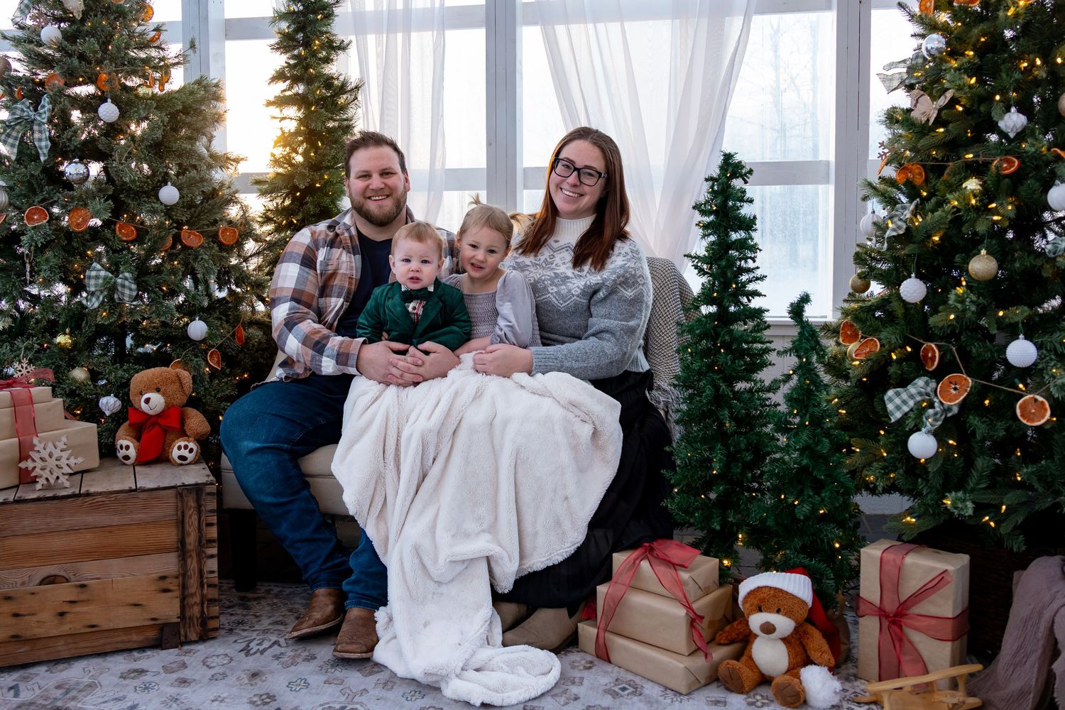 Family of four posing for Christmas portrait surrounded by decorated trees and gifts.