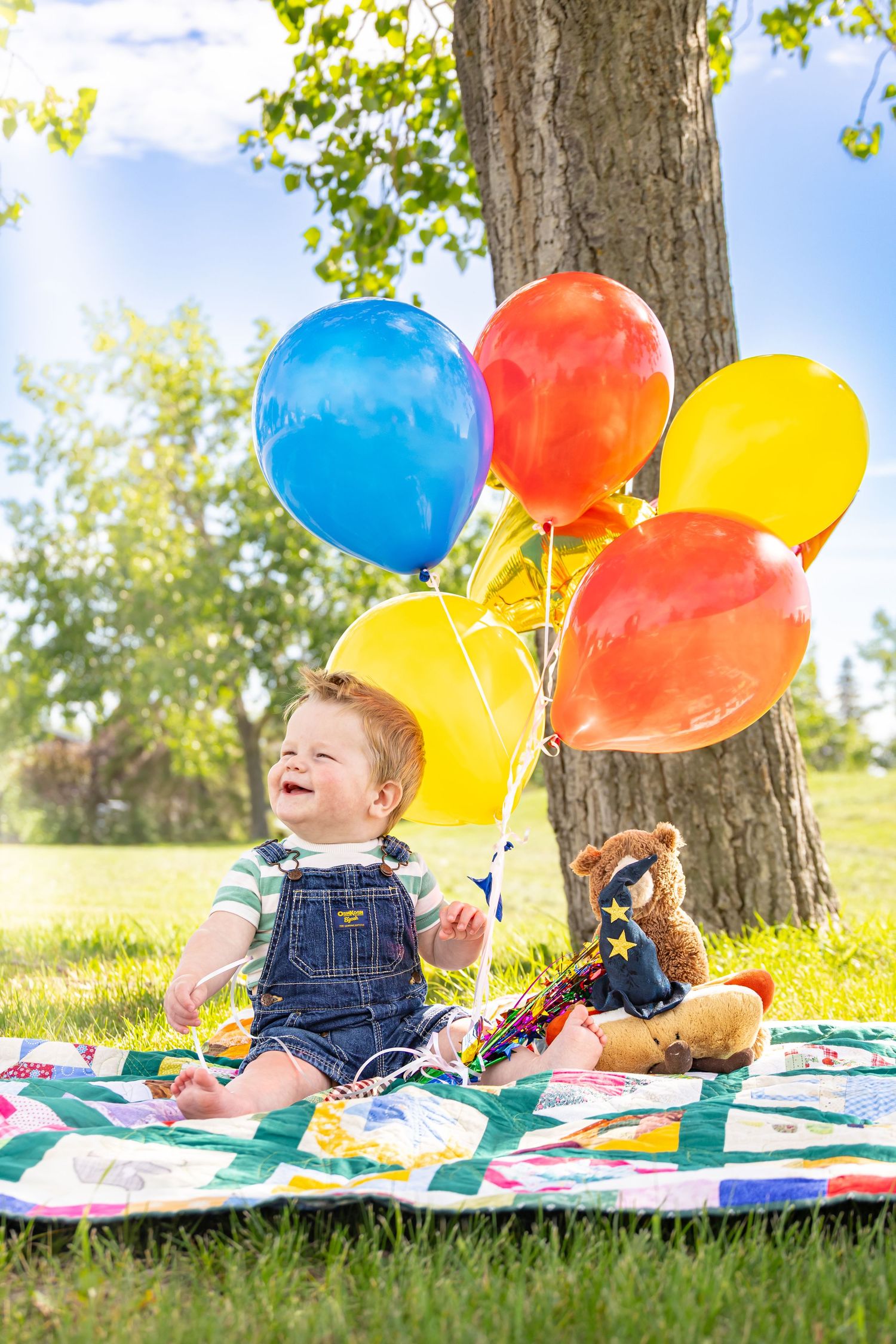Happy toddler in denim overalls sits on a quilt holding colorful balloons beside a teddy bear in a sunny park.