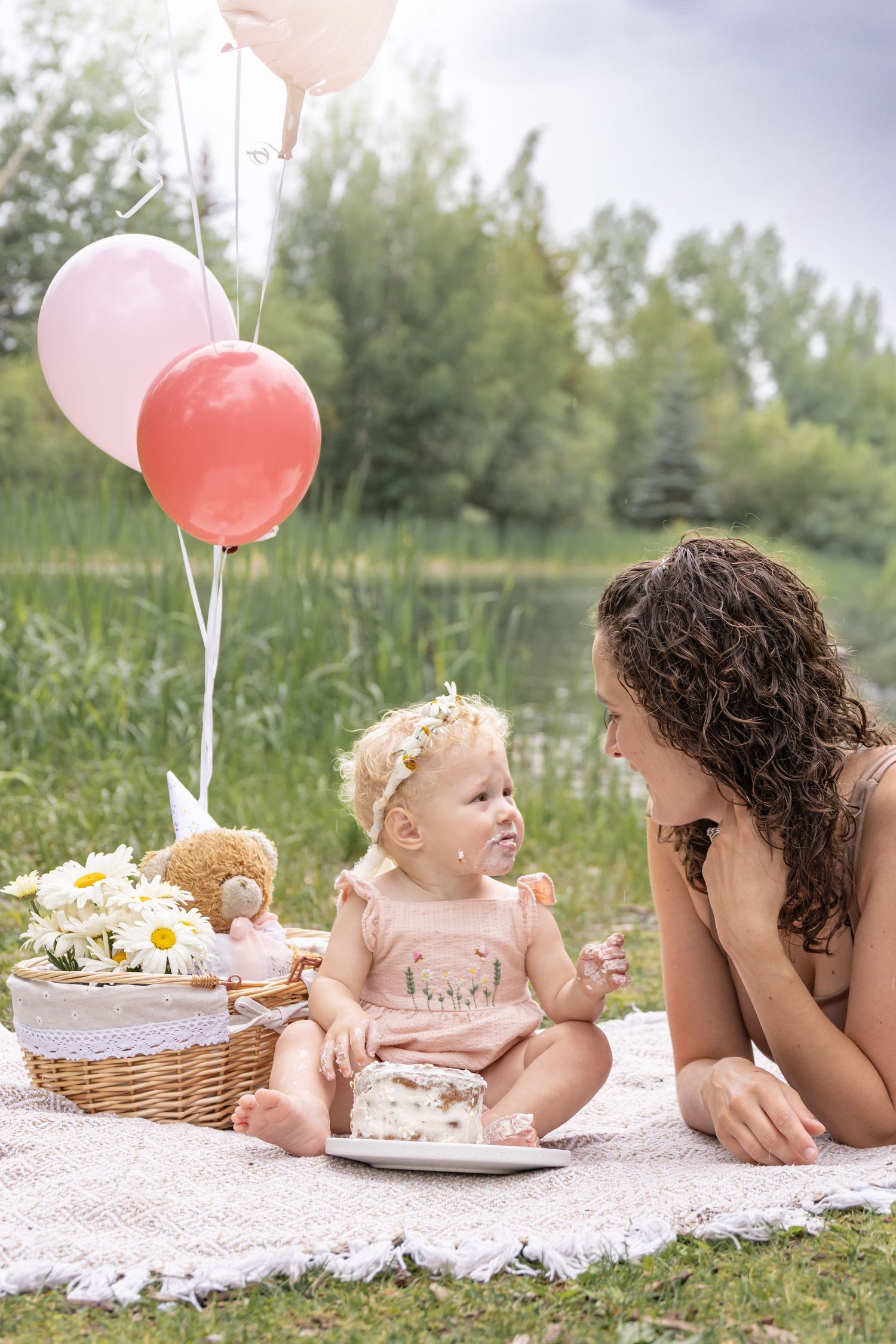 Baby in a flower crown enjoying a smash cake on a picnic blanket beside a smiling mother, with balloons and a daisy basket by a lake.