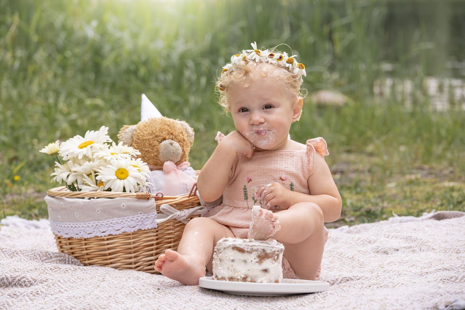 Toddler in a floral crown sits on a blanket in a sunny meadow, enjoying a small cake beside a daisy basket and teddy bear.