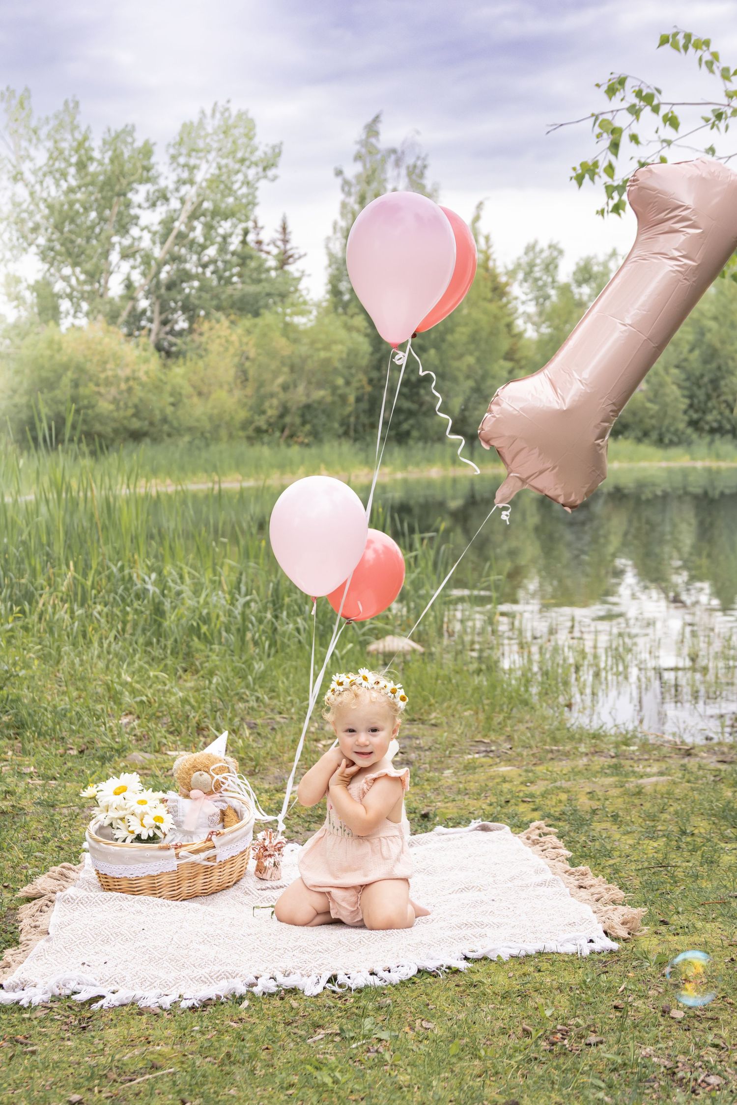 Baby girl sits on blanket outdoors with pink balloons and number 1 balloon celebrating her first birthday by a pond.