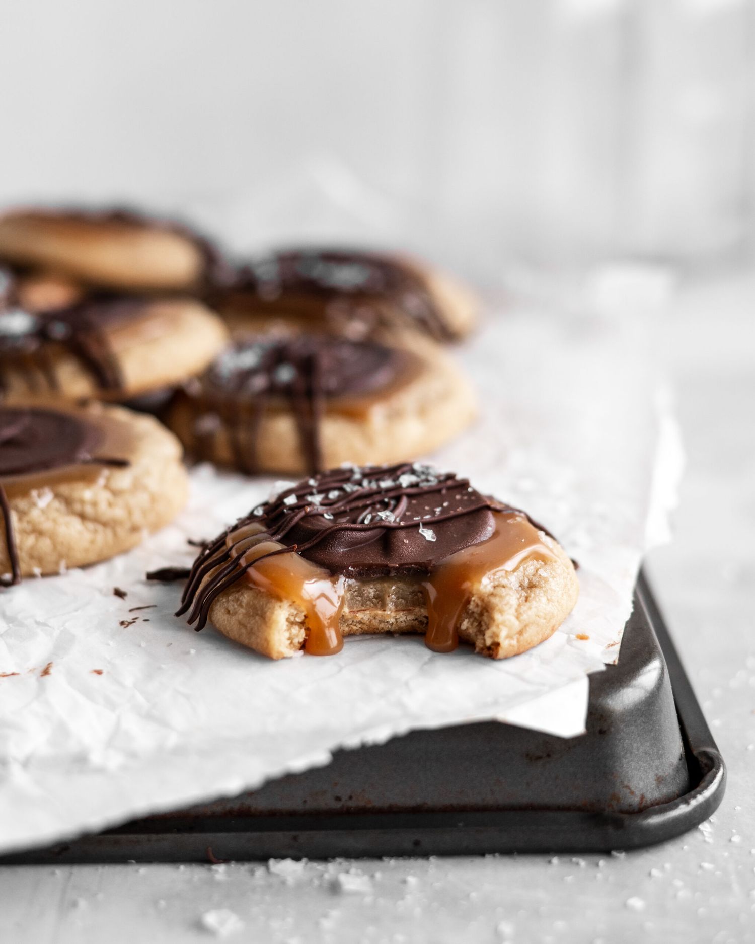 Chocolate and caramel dipped shortbread cookies with bite taken out revealing gooey center on dark baking sheet.