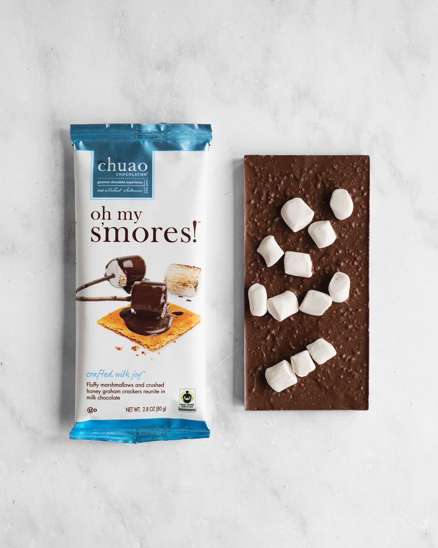Overhead view of chocolate covered graham crackers with marshmallows arranged on a white marble surface with product packaging.