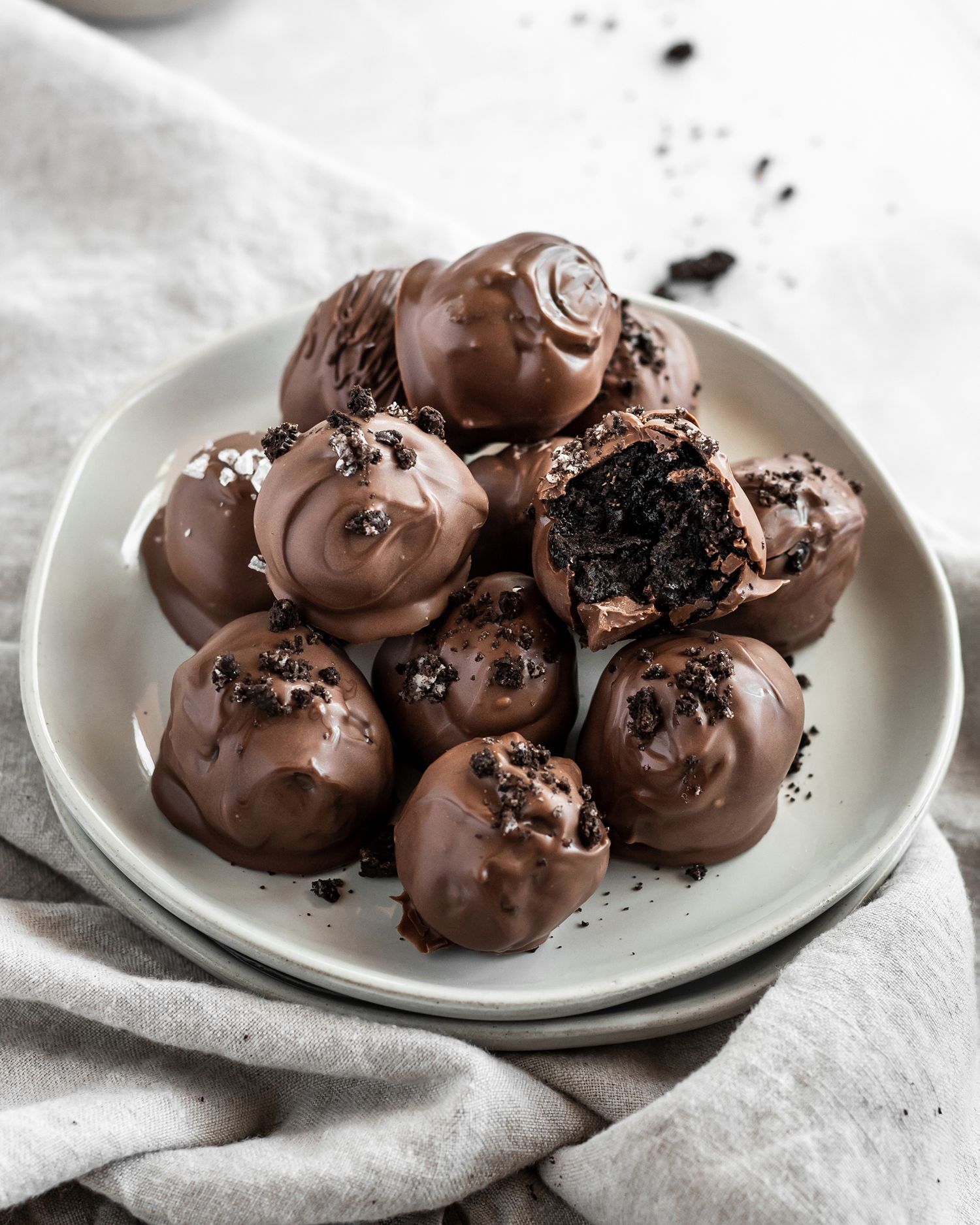Chocolate-coated Oreo truffles garnished with chocolate sprinkles arranged on a white plate with a linen napkin.