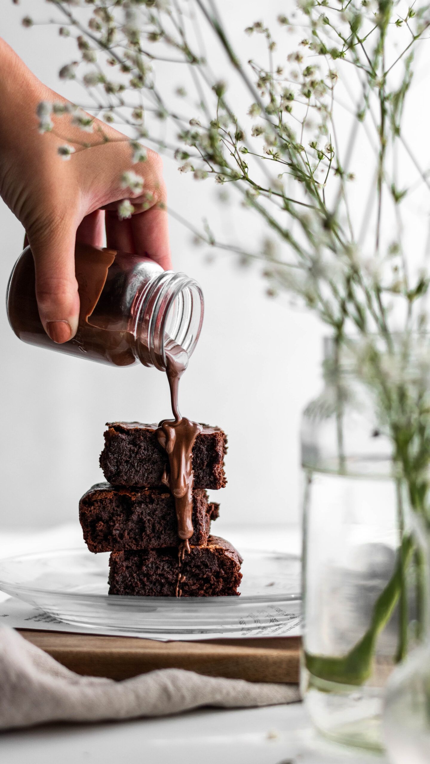 Chocolate sauce being poured over a stack of rich brownies with delicate white flowers in the background.