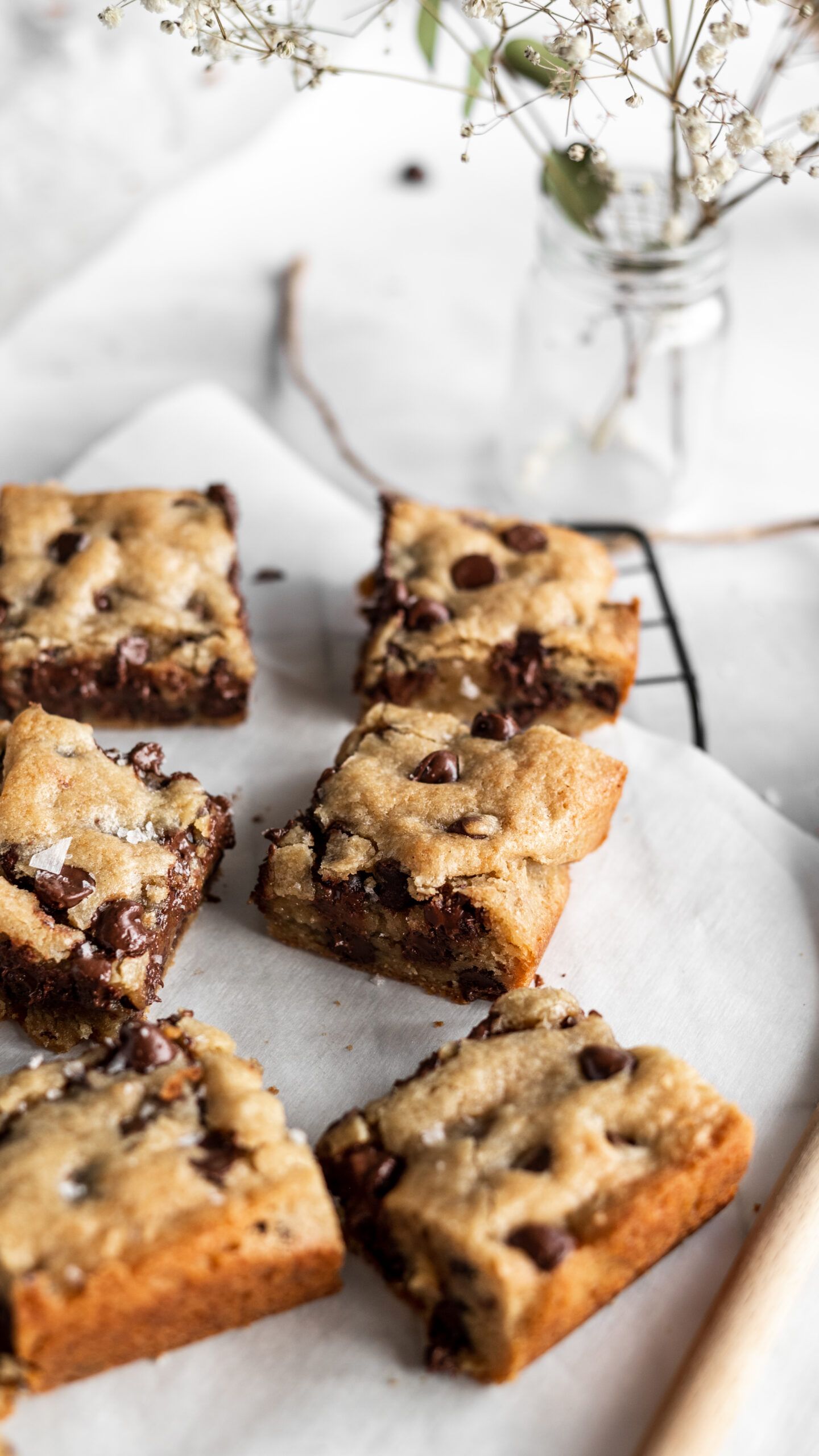 Homemade chocolate chip cookie bars displayed on parchment paper with delicate white flowers in a vase in the background.