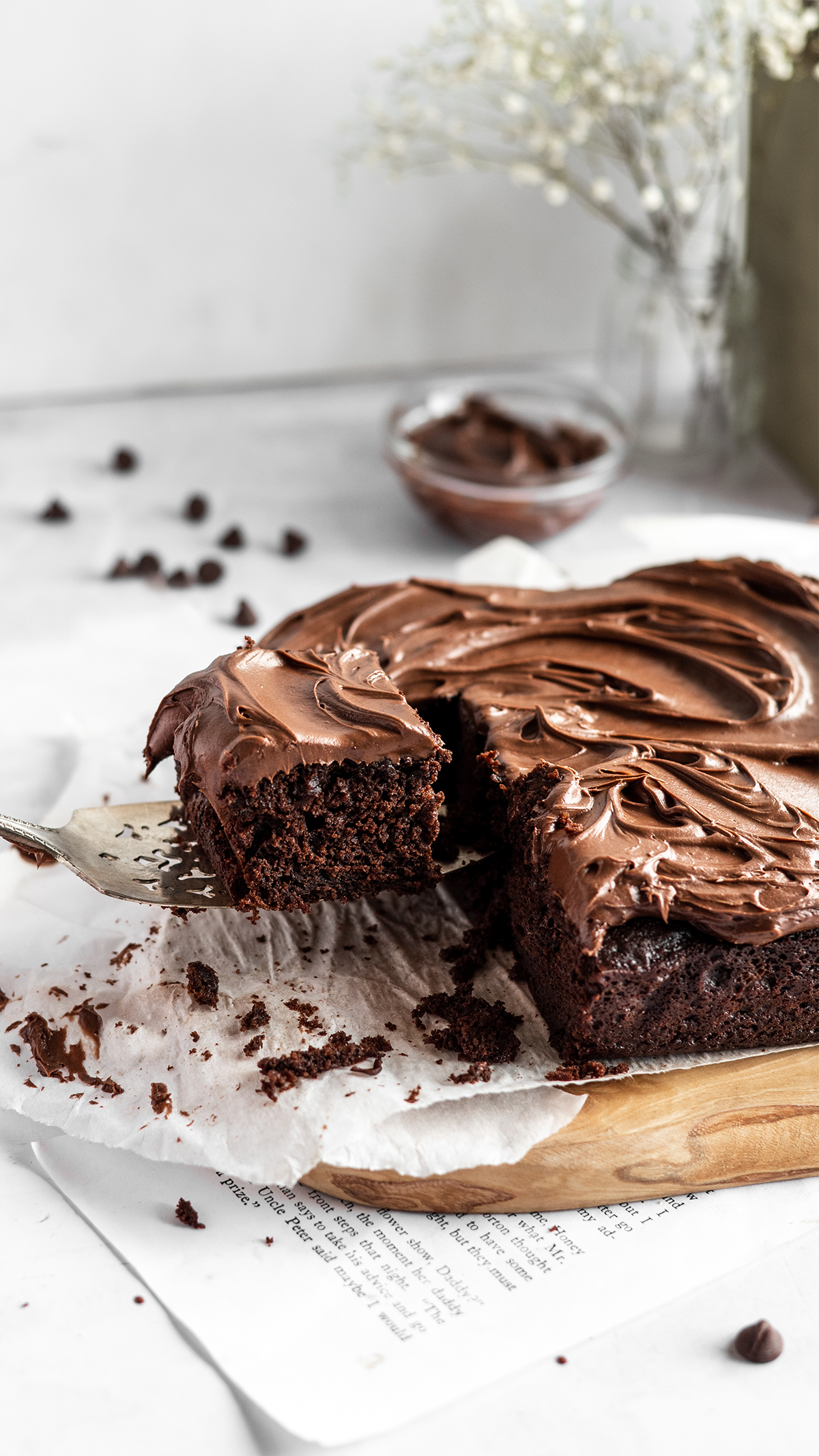 Homemade chocolate brownie with rich chocolate frosting on a white parchment paper with chocolate chips scattered around.