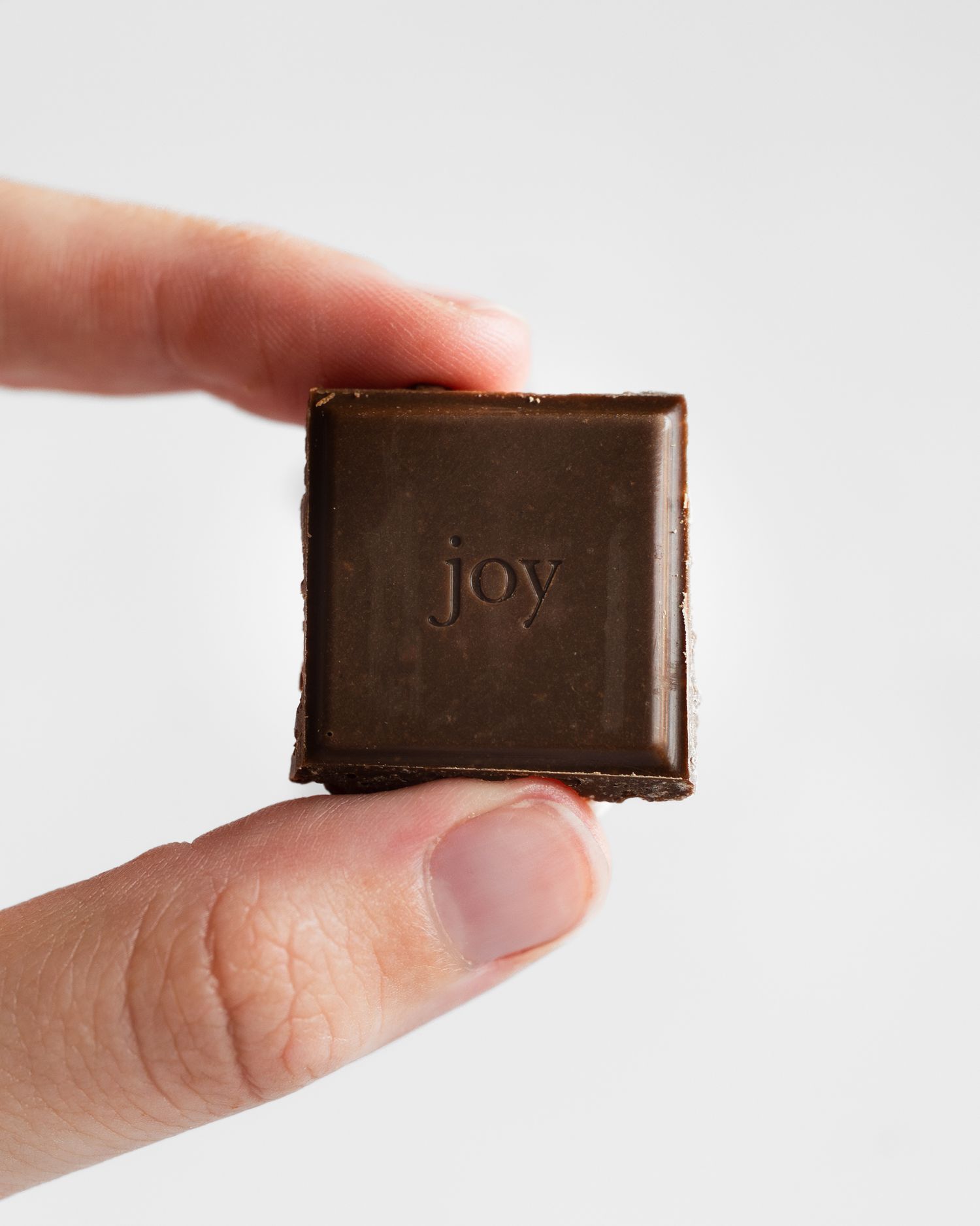 Close-up of a small square piece of dark chocolate with the word 'joy' embossed on its surface.