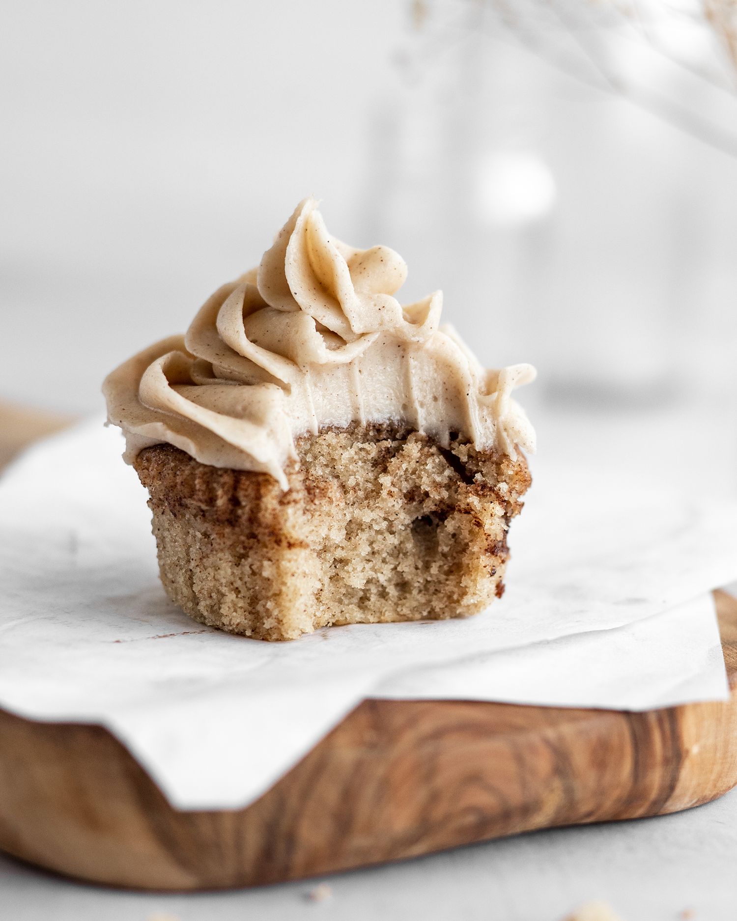 A bitten vanilla cupcake with creamy brown frosting sits on a white napkin over a wooden serving board.