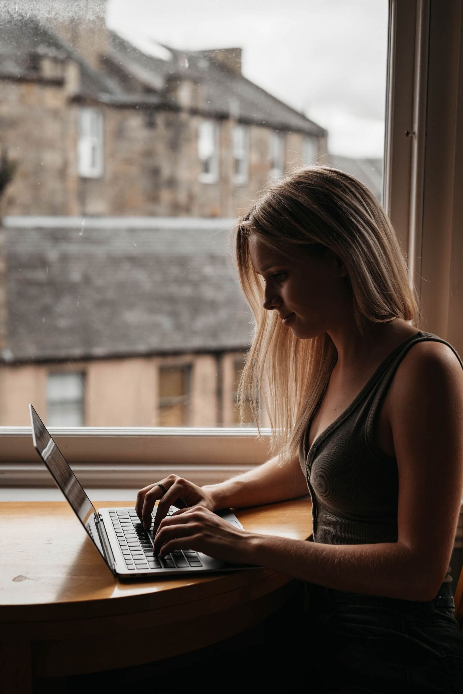 Someone works on a laptop computer by a window with moody natural lighting and old buildings visible outside.