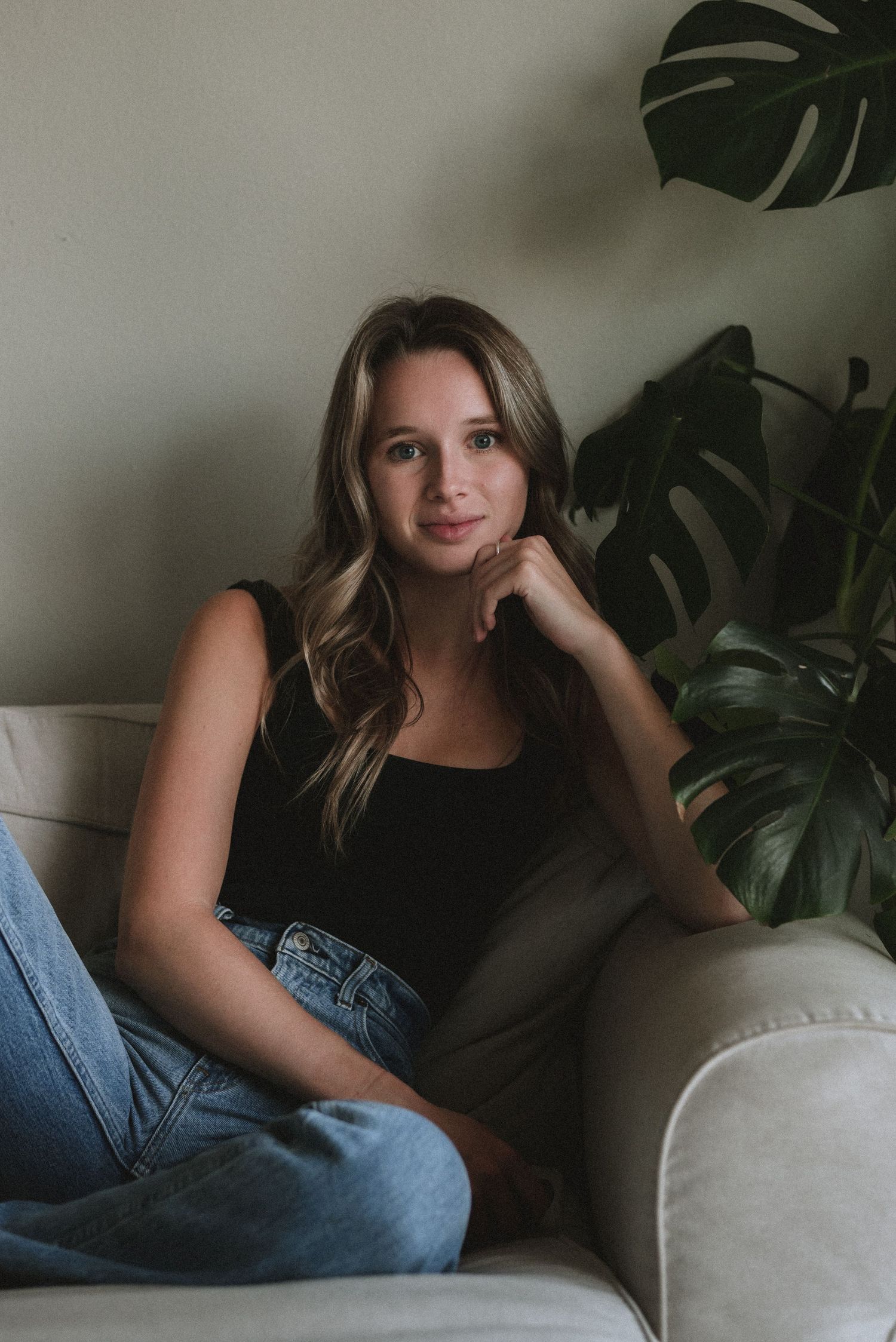 Portrait of someone relaxing on a couch in a black top and jeans with monstera plant leaves in the background.