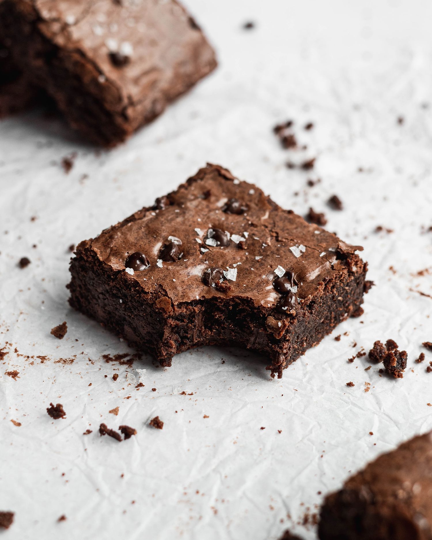A rich chocolate brownie with a fudgy texture and crumbly edges sits on a white surface with a bite missing.