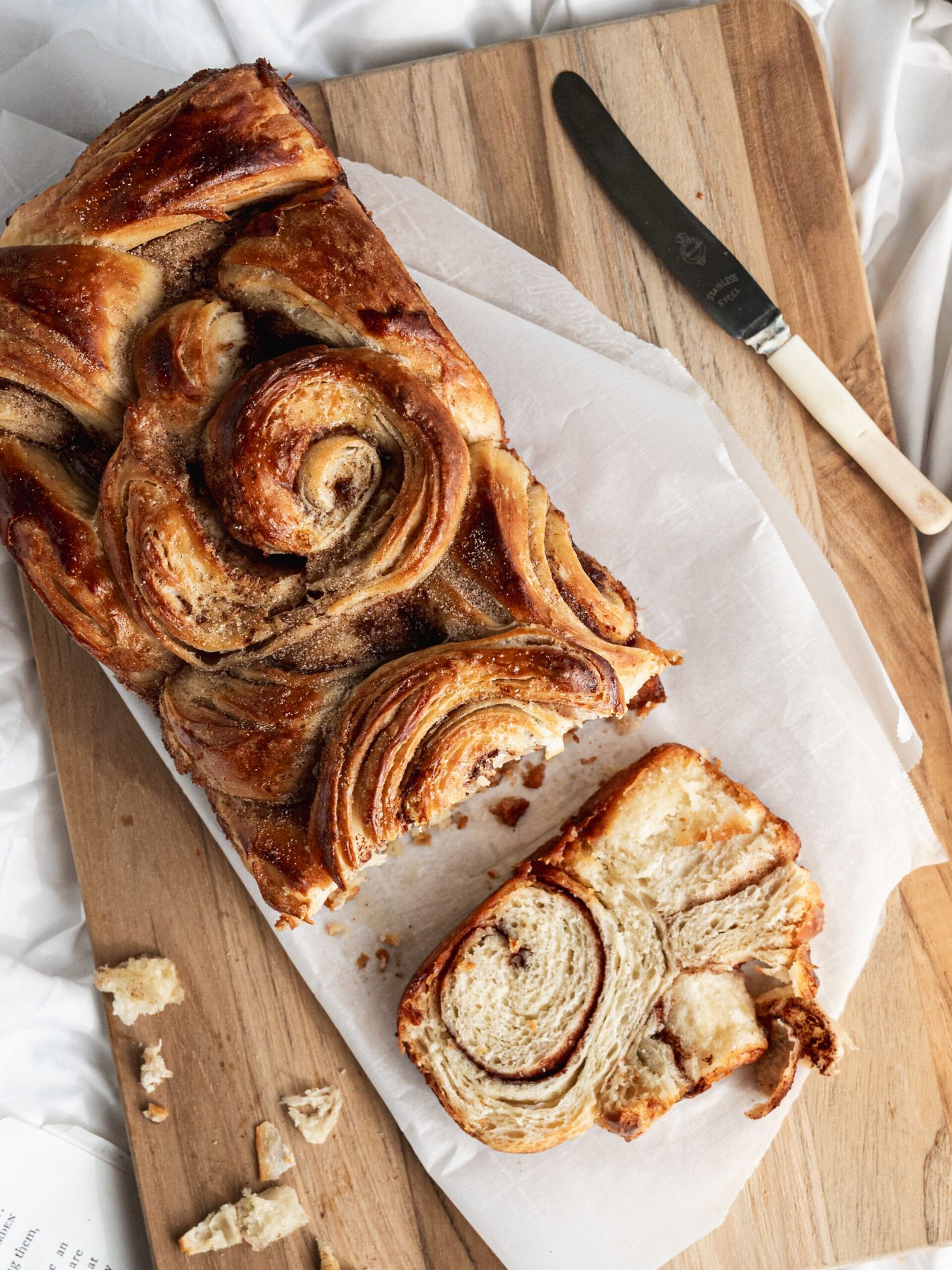 Homemade cinnamon swirl babka bread loaf with flaky layers on wooden cutting board with knife and parchment paper.