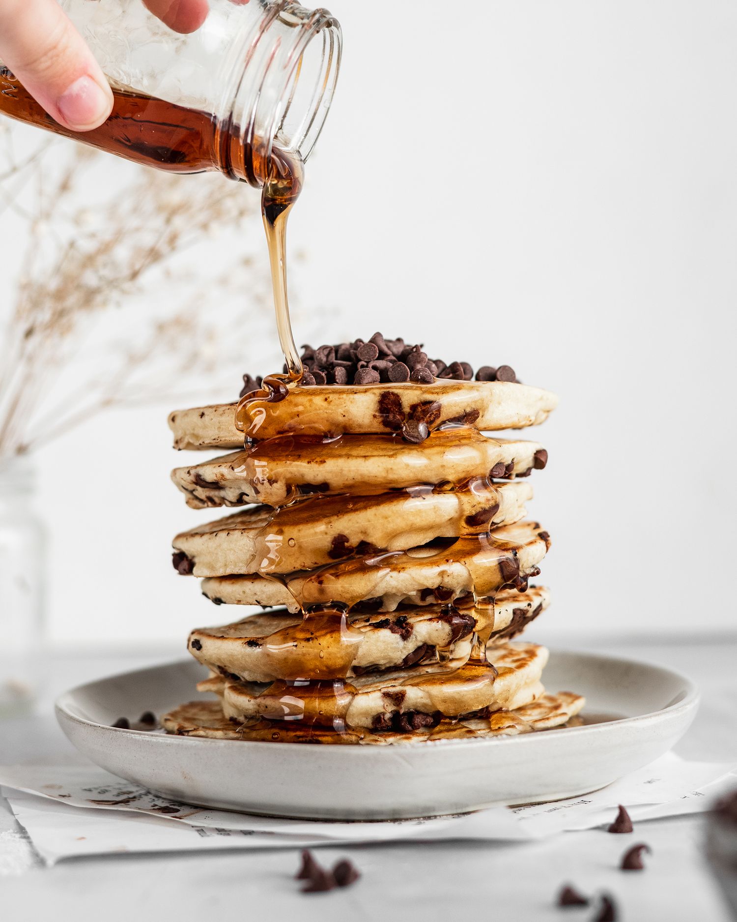 Chocolate chip pancakes stacked high on a white plate with maple syrup being drizzled from a glass bottle.
