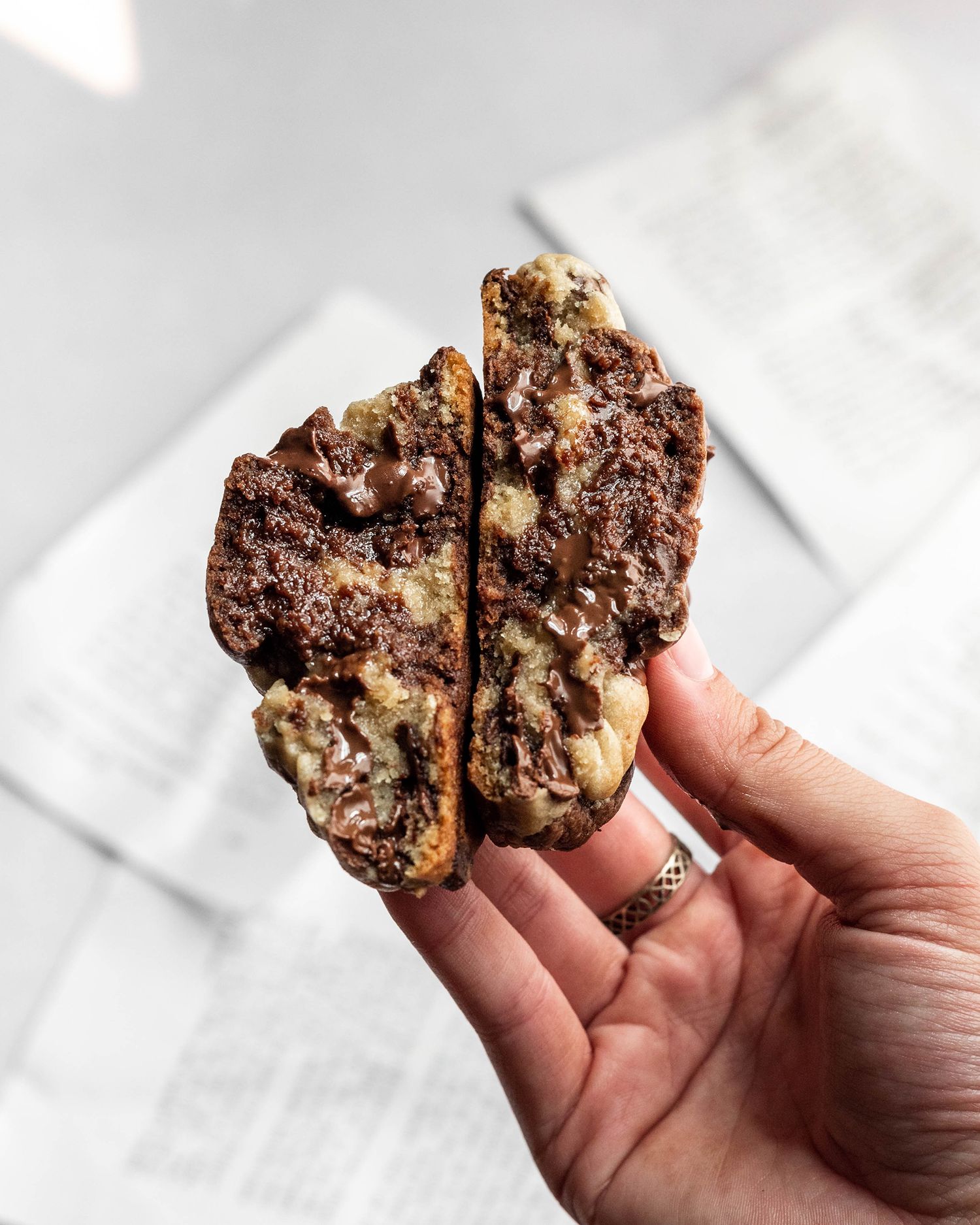 Hand holding a broken chocolate chip cookie revealing a soft and marbled interior texture on a light background.