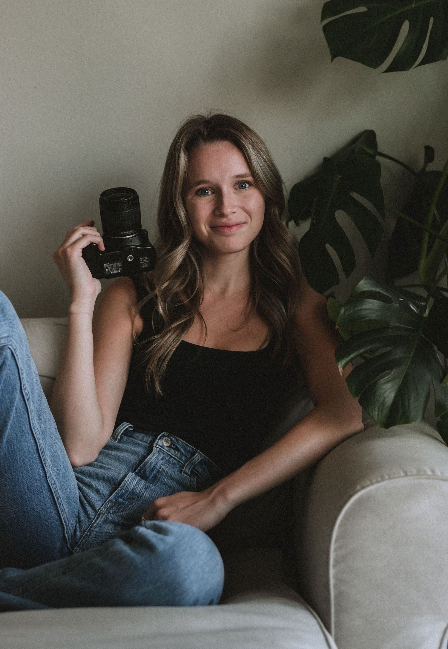 A person in casual black top and jeans relaxes on a gray couch with monstera plants in the background.