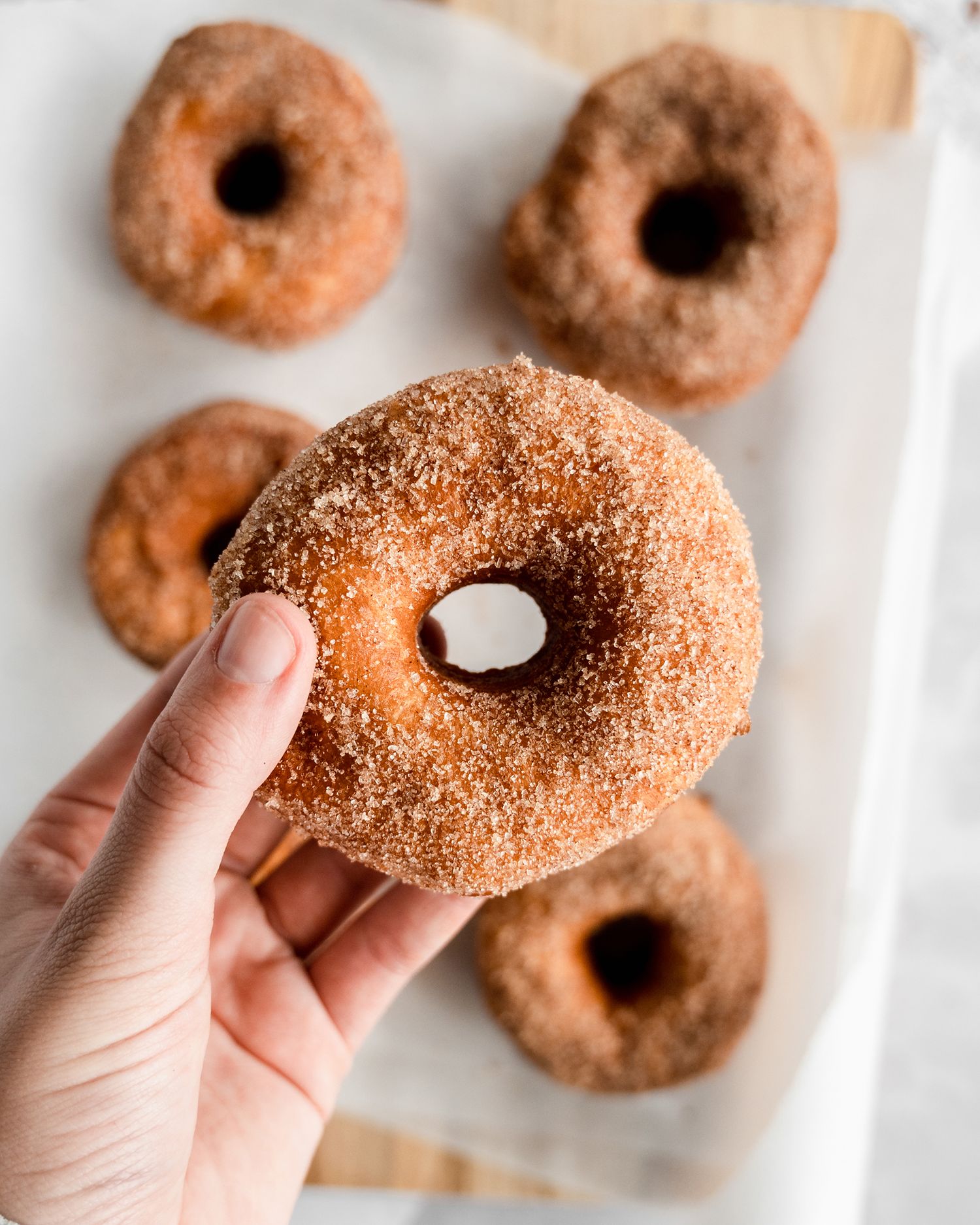 Hand holding a freshly made apple cider donut coated in cinnamon sugar on white parchment paper.
