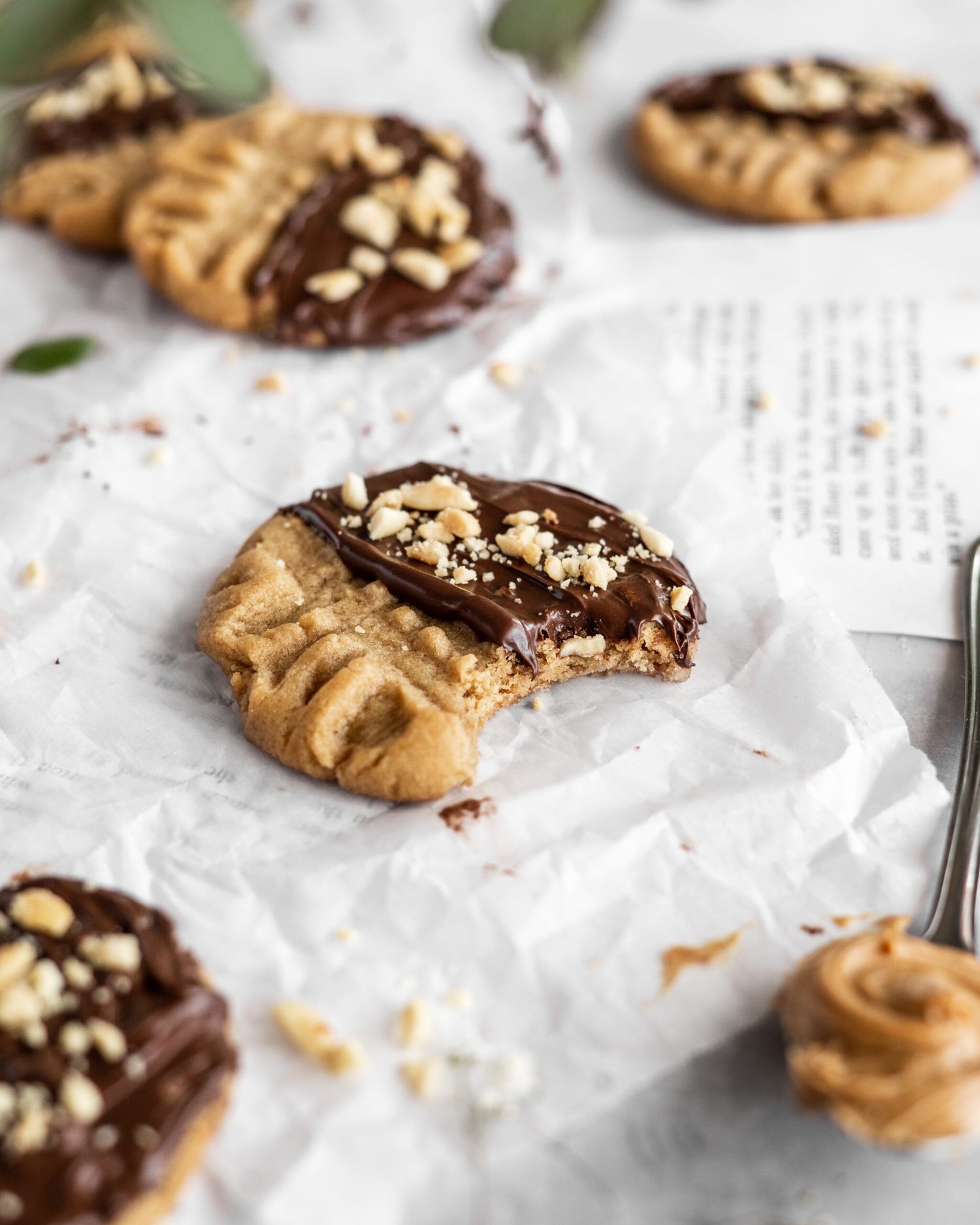 Chocolate-dipped peanut butter cookies with chopped nuts scattered on white parchment paper.