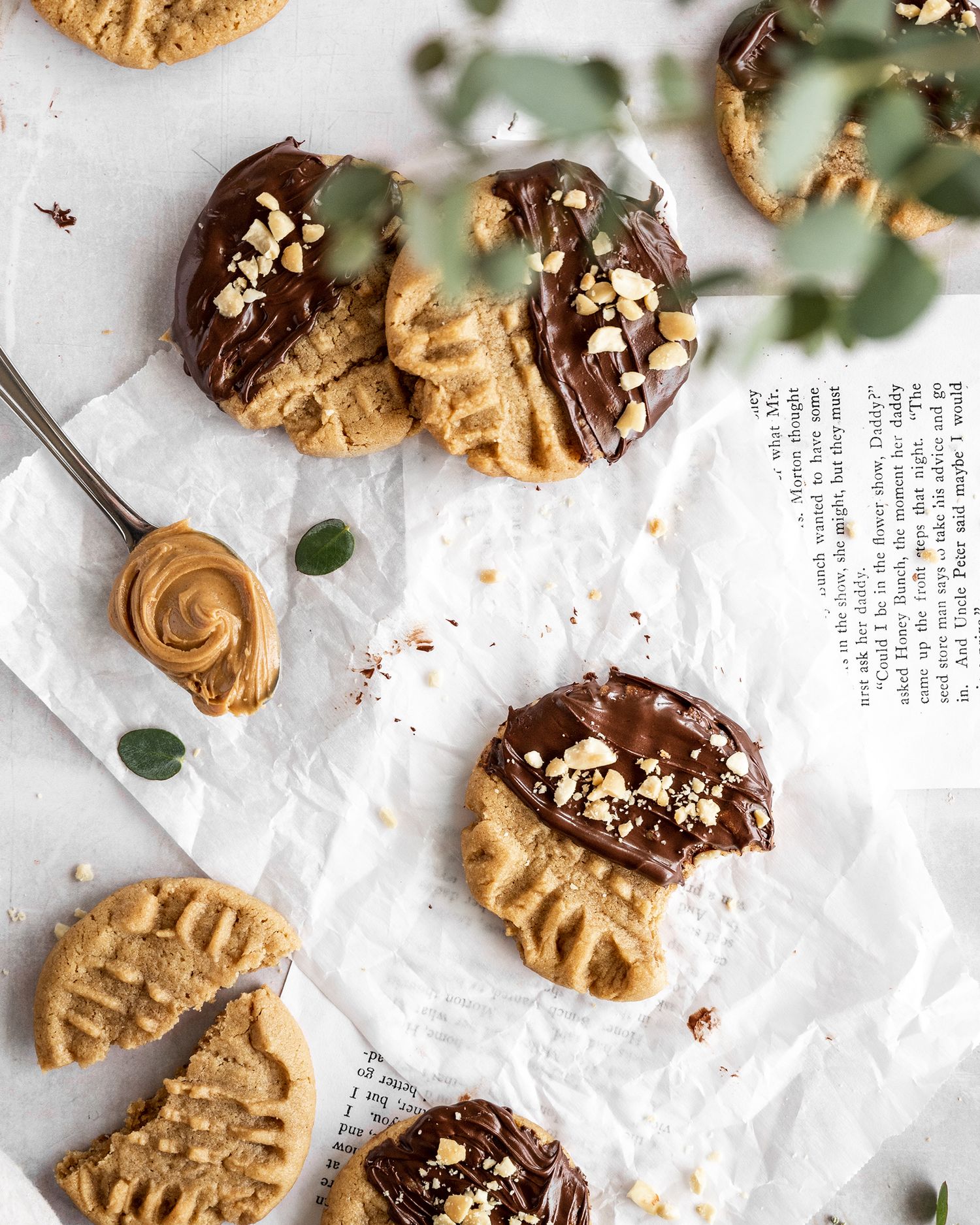 Homemade chocolate-dipped peanut butter cookies with chopped nuts scattered on white parchment paper next to a jar of peanut butter.