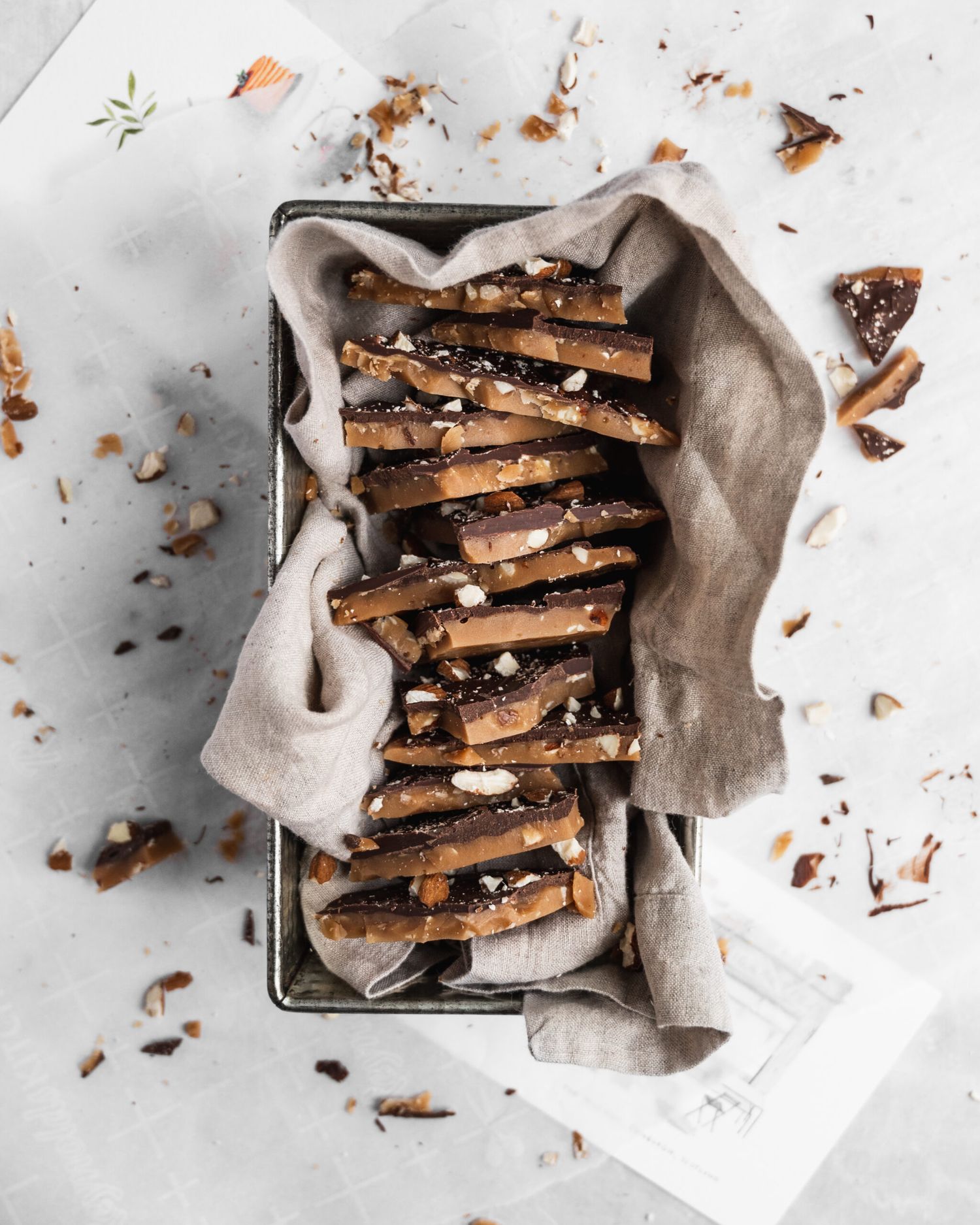 Stacked chocolate toffee bars with crumbs scattered on a grey linen napkin in a rustic metal baking pan.