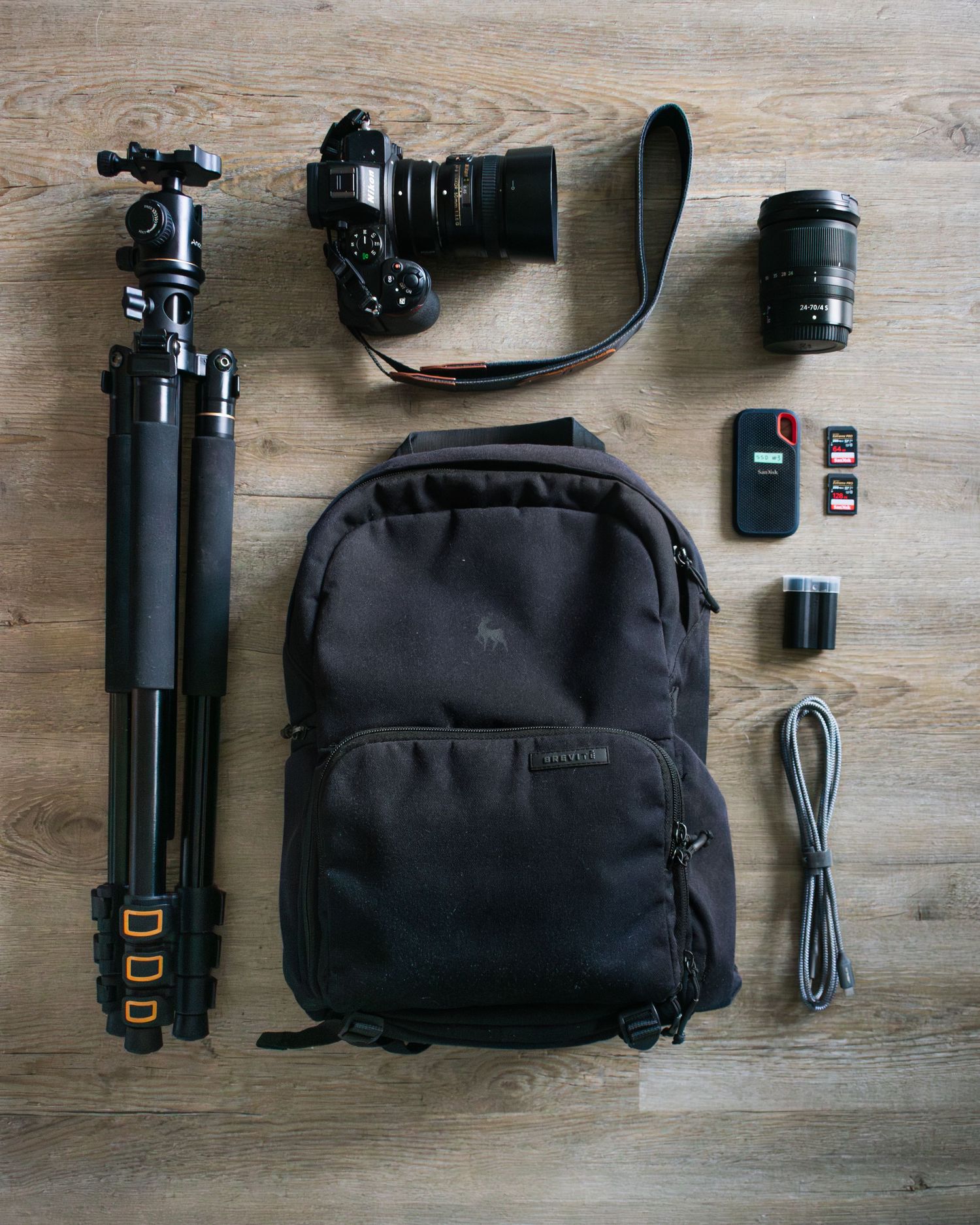 Flat lay of photography gear including camera, tripod, backpack, lenses and accessories on wooden surface.